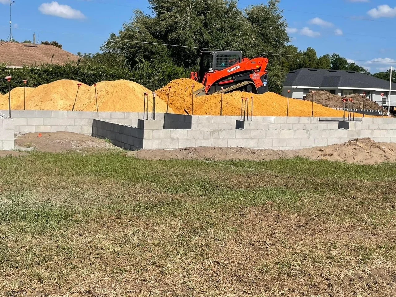Construction site with cinder block foundation walls, piles of yellow and orange sand, and a small orange Kubota skid steer loader on top of the sand. Residential houses in the background under partly cloudy sky.