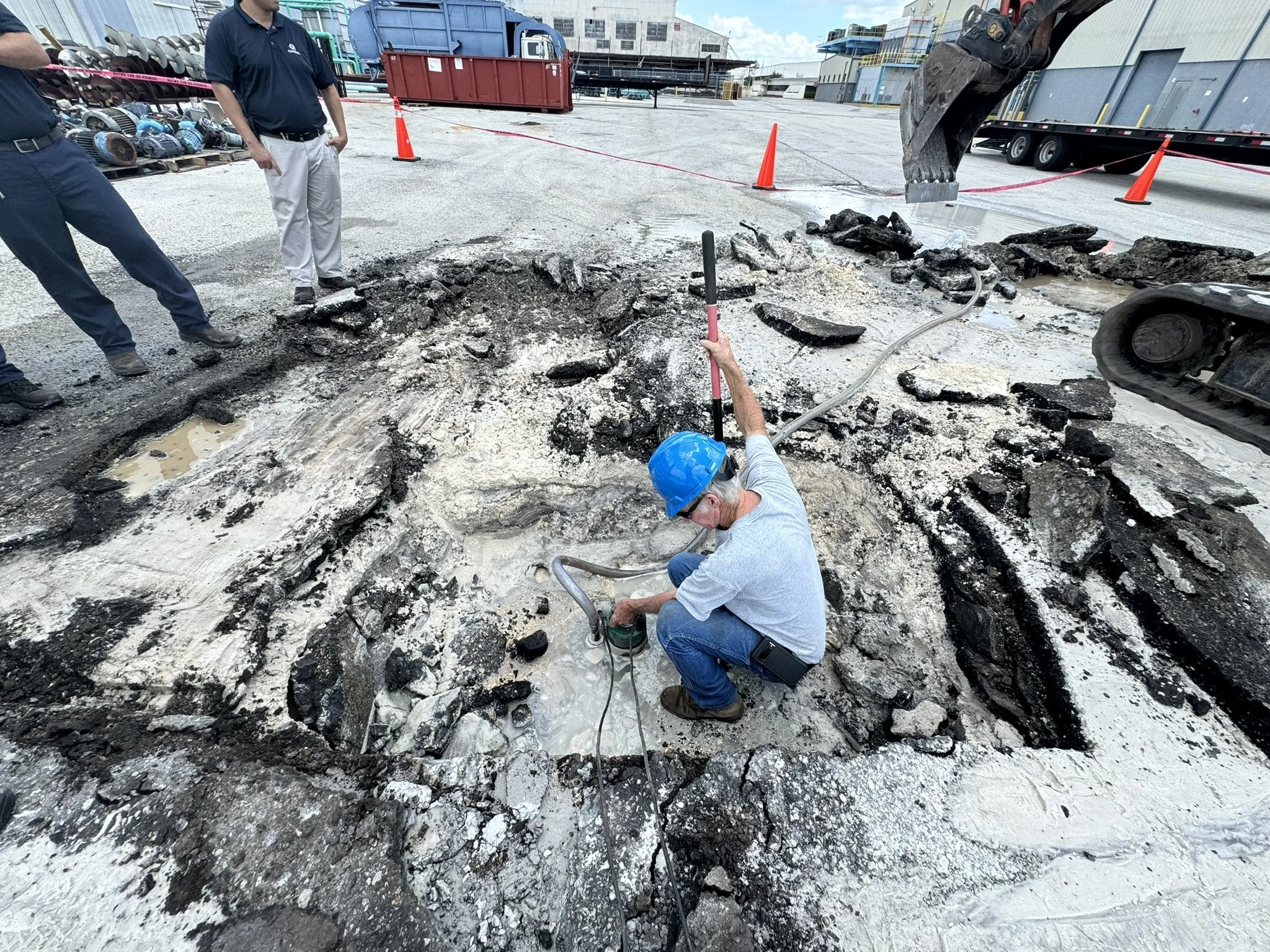 Construction workers examining a large hole in the ground with asphalt and rubble around it, near construction equipment and trailers.