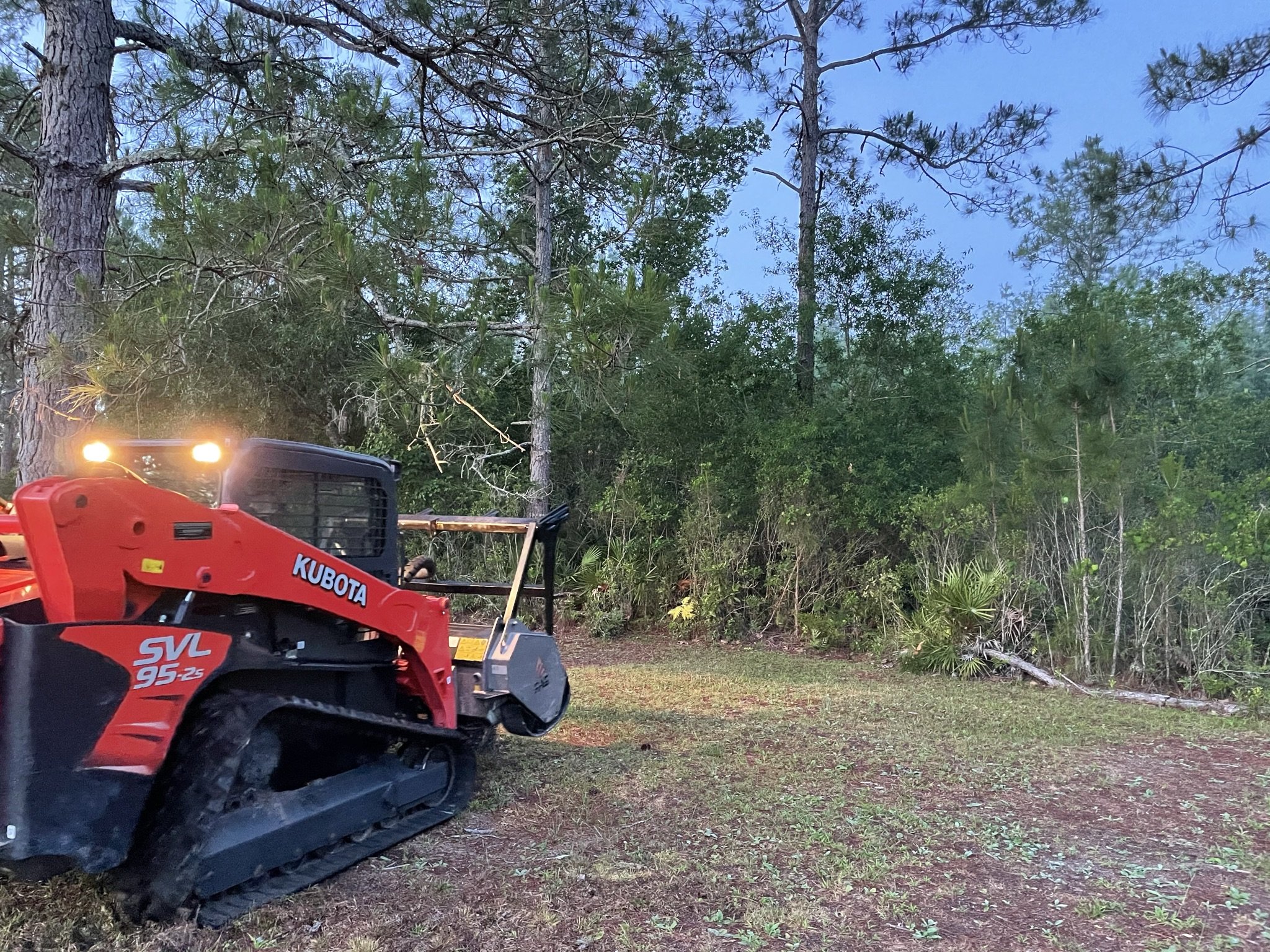 Close-up of a red Kubota SVL95-2S compact track loader on a grassy area in front of trees and bushes, with the vehicle's front facing left and the rear with bright headlights.