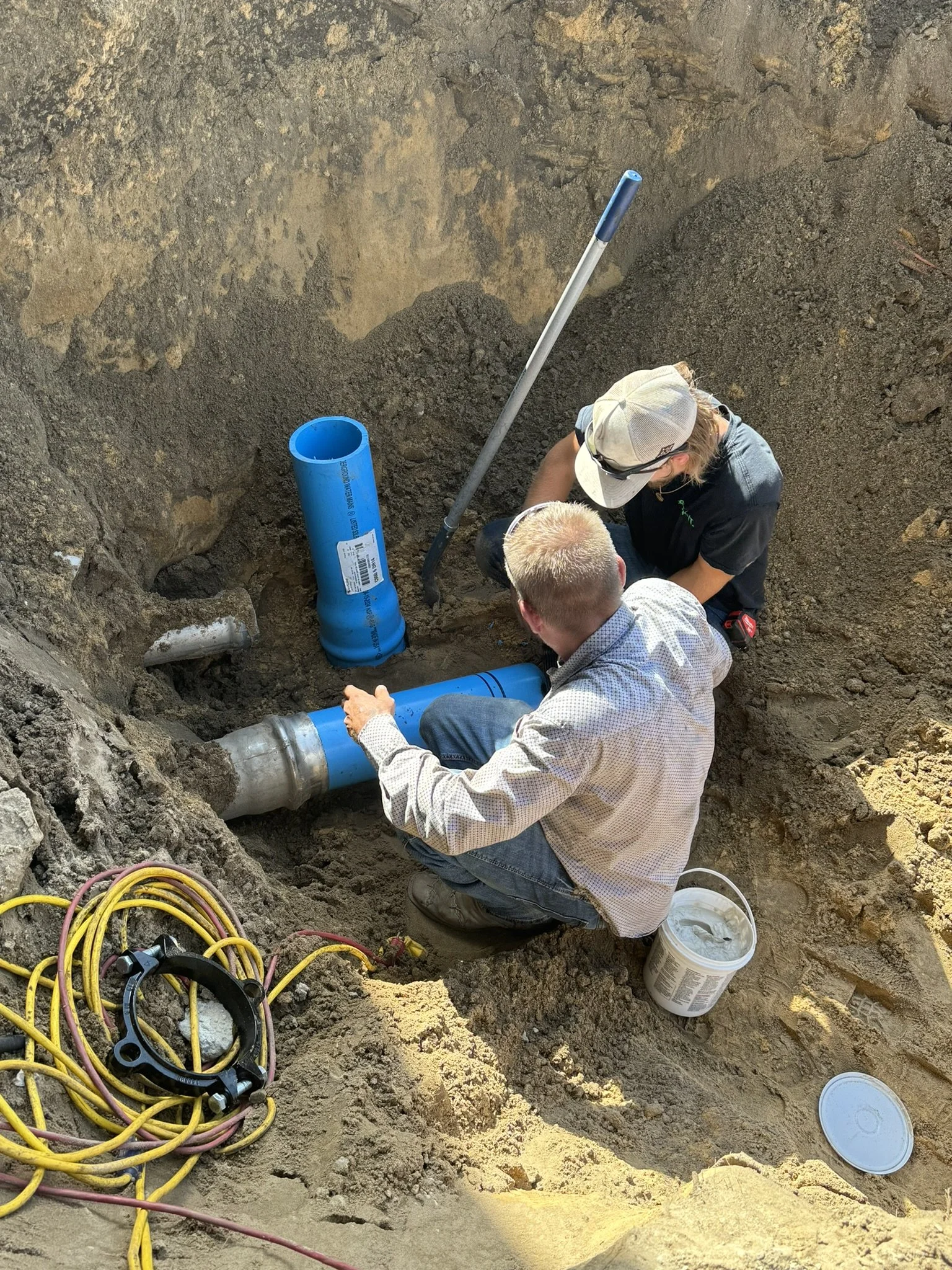 Two workers are installing underground piping in a dirt excavation site. One man wearing a cap and sunglasses is connecting a large blue pipe to a metal pipe, while another man observes. Several coiled electrical cords and tools are on the ground nearby.