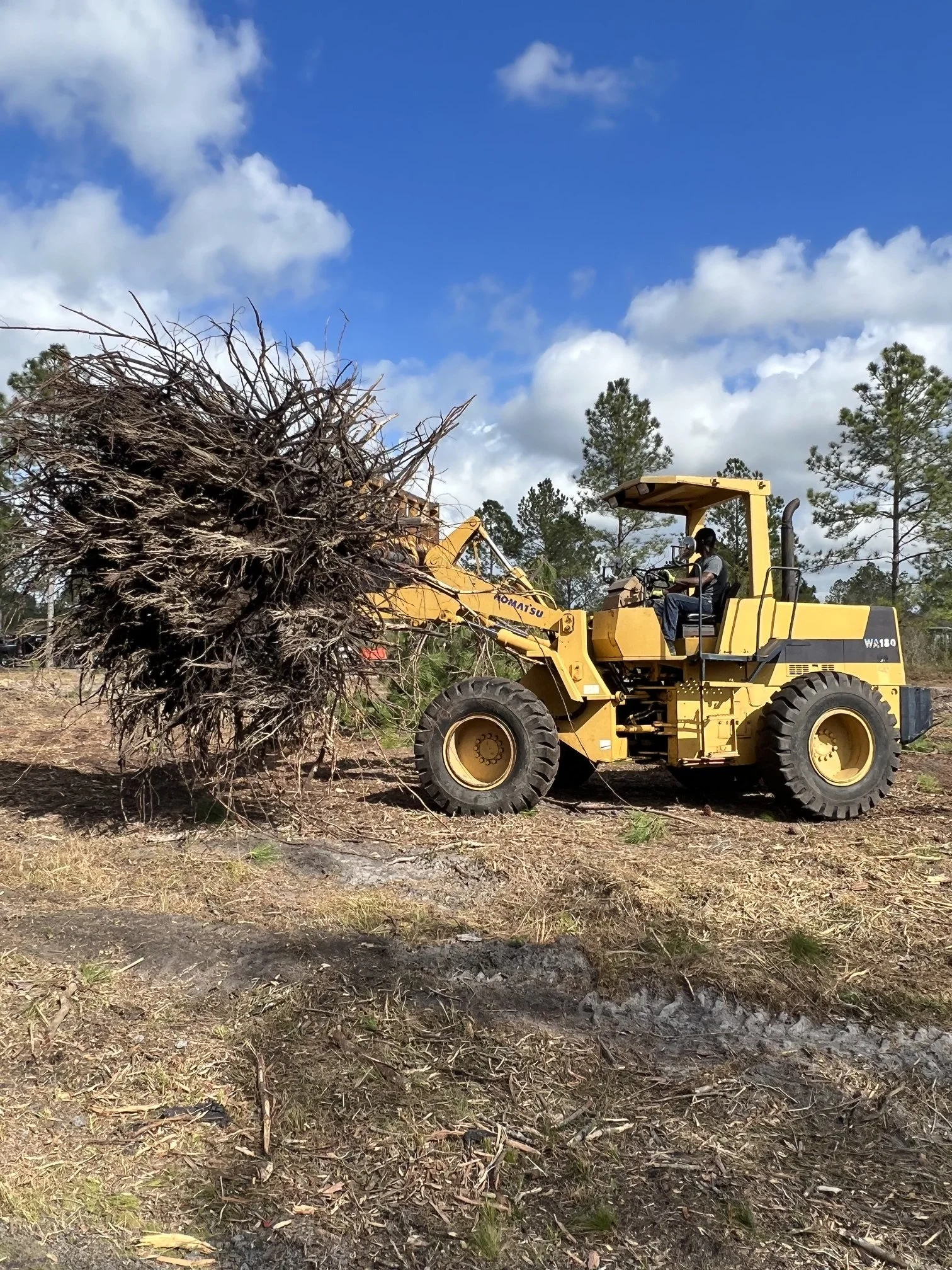 A construction worker operating a yellow bulldozer on a cleared land with trees and a partly cloudy sky in the background.