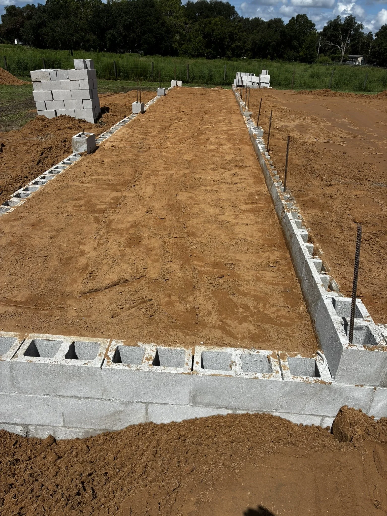 Construction site with cinder block foundation and a dirt pathway, surrounded by trees and open land.