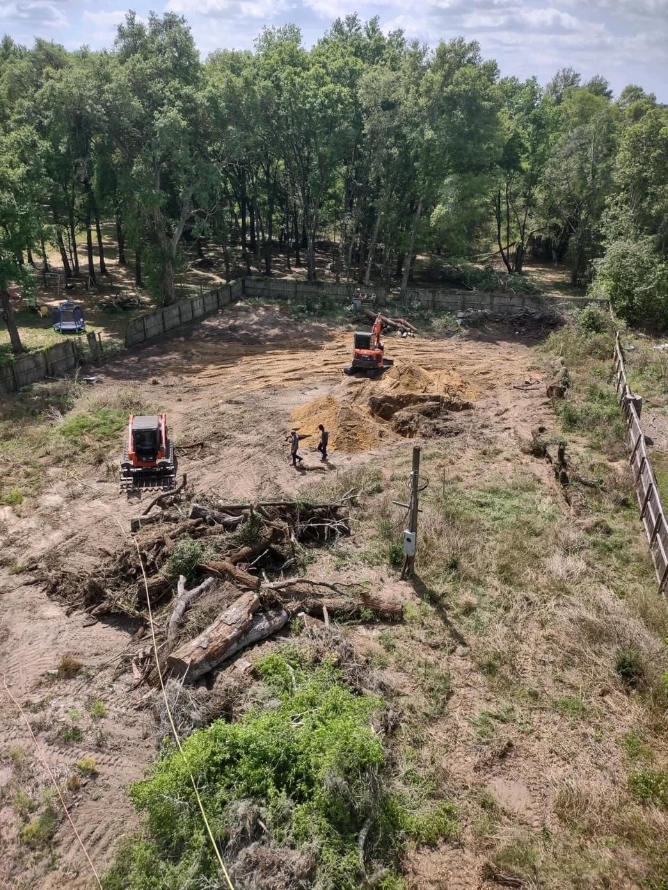 A construction site with two excavators, one digging and one stationary, and two workers standing and talking. The site is surrounded by trees and a wooden fence, and there are logs and debris in the foreground.