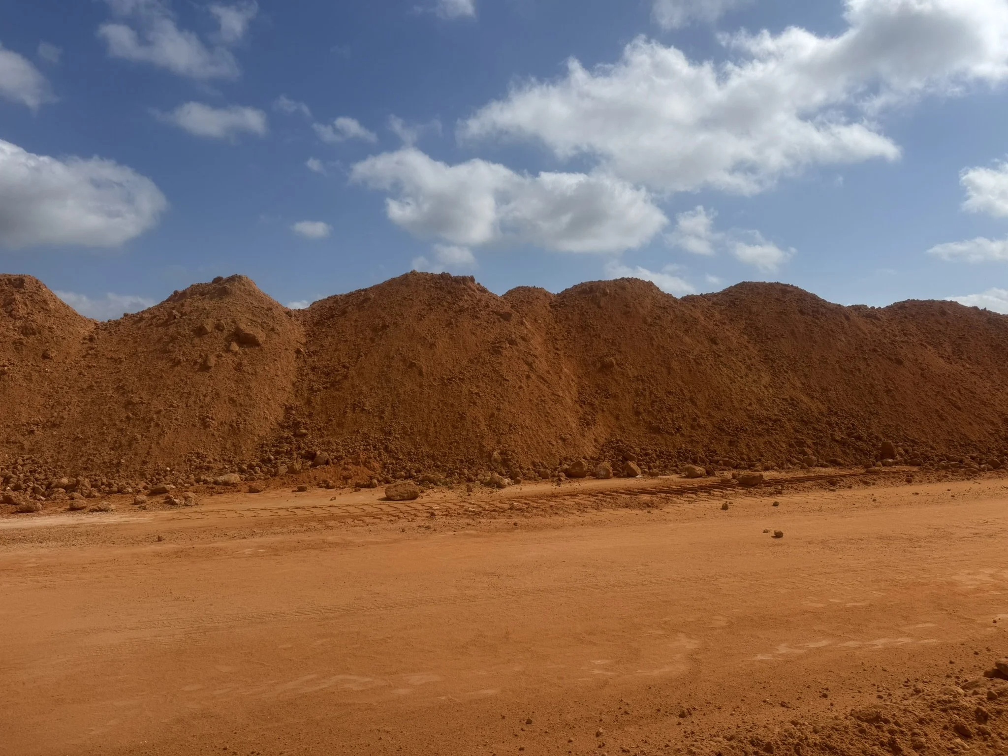 Bright blue sky with scattered white clouds above reddish-brown desert terrain with rugged hills in the background.