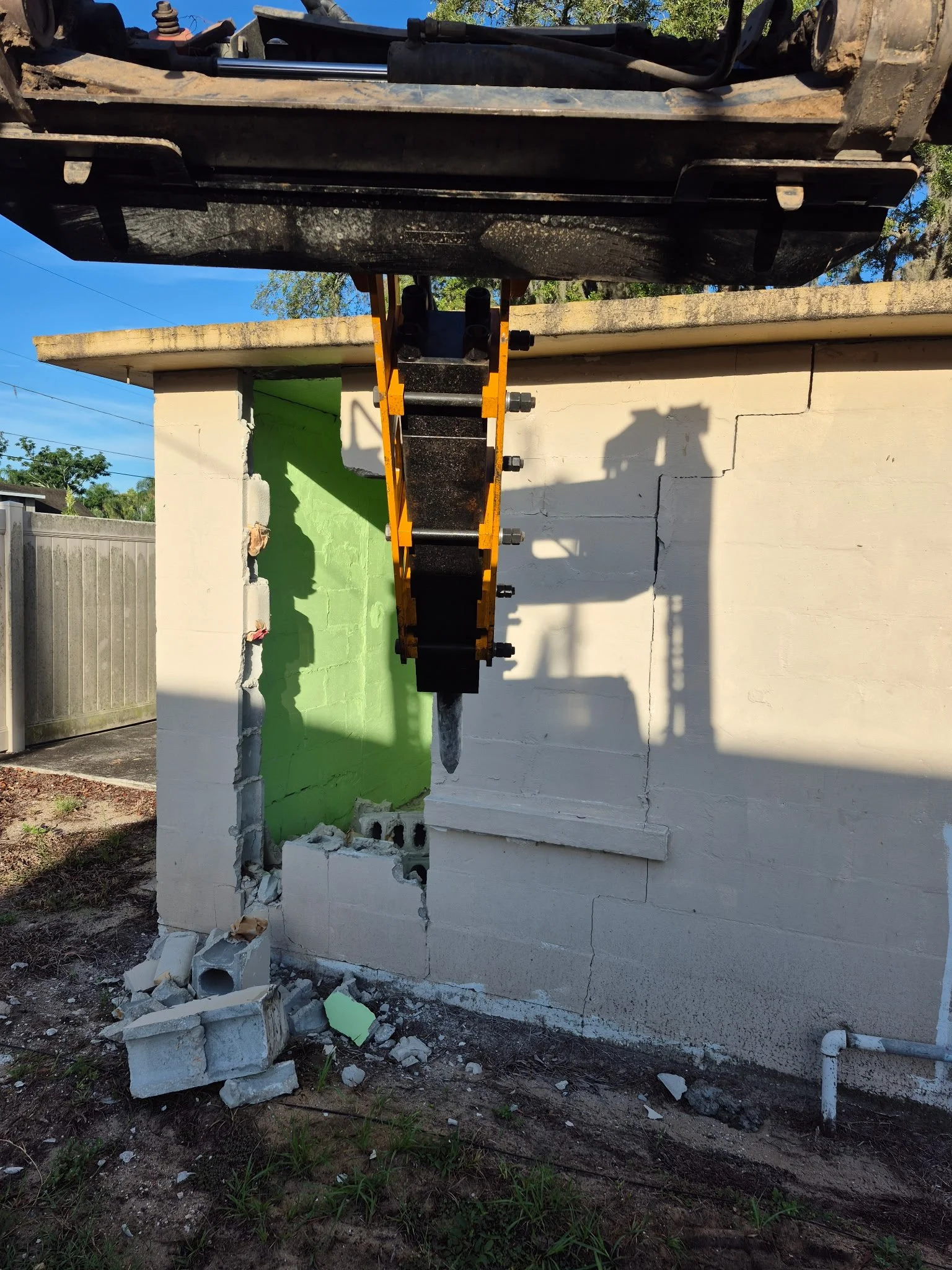 The image shows a construction site where part of a building wall is being demolished or repaired. There is a hydraulic breaker attached to a piece of heavy machinery, with debris and broken concrete at the base of the wall. The wall has a small section painted green, and a shadow of the machinery is cast on the wall.