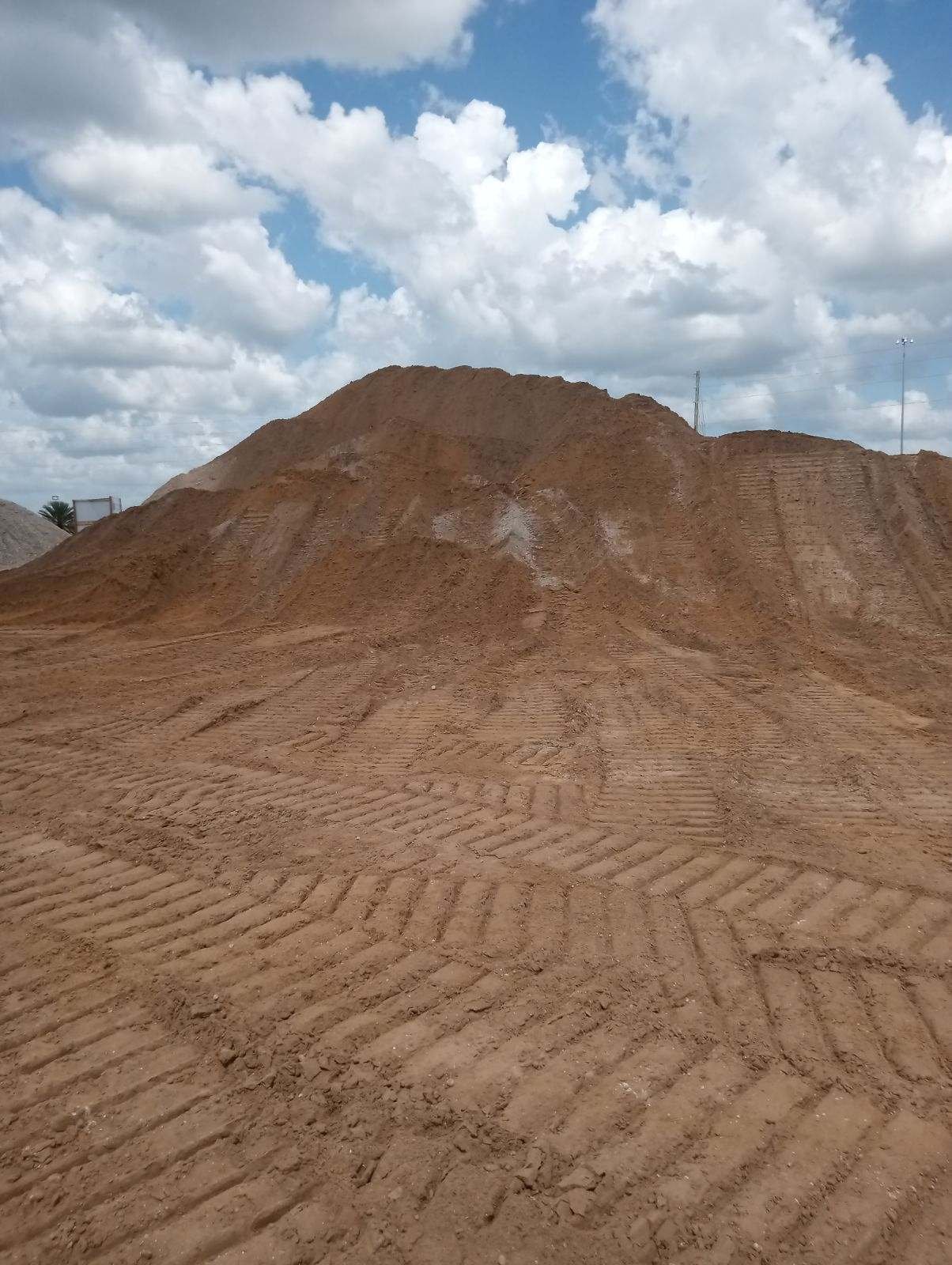 A large dirt pile or mound on a construction site with tire tracks in the foreground and a partly cloudy sky above.