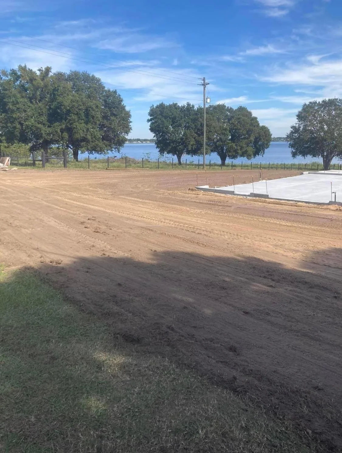 Construction site with a dirt road, grassy area, and a concrete foundation under construction, near trees and a body of water under a partly cloudy sky.