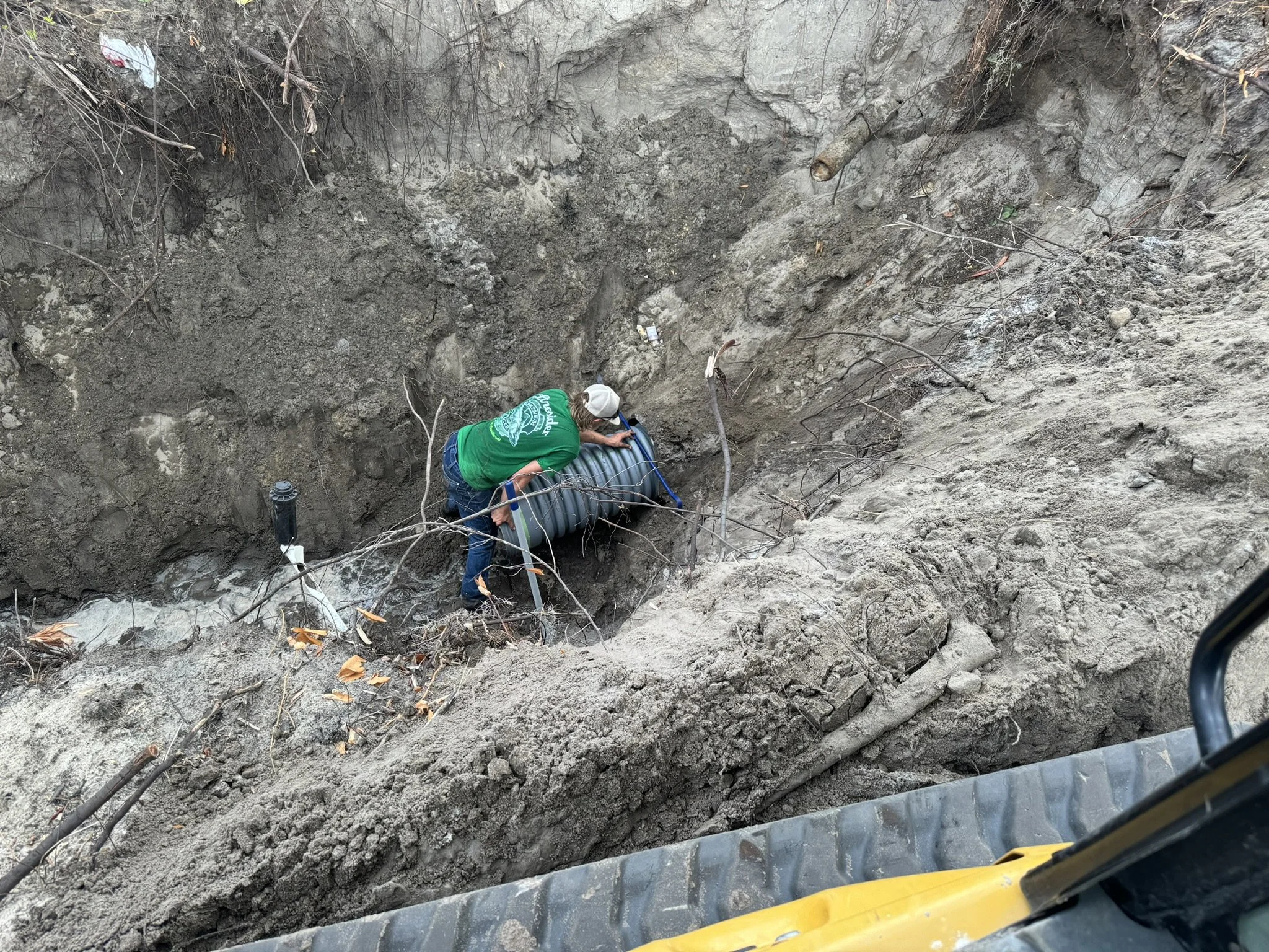 Worker in green shirt and white helmet installing or repairing a large underground pipe in a deep trench surrounded by dirt and soil.