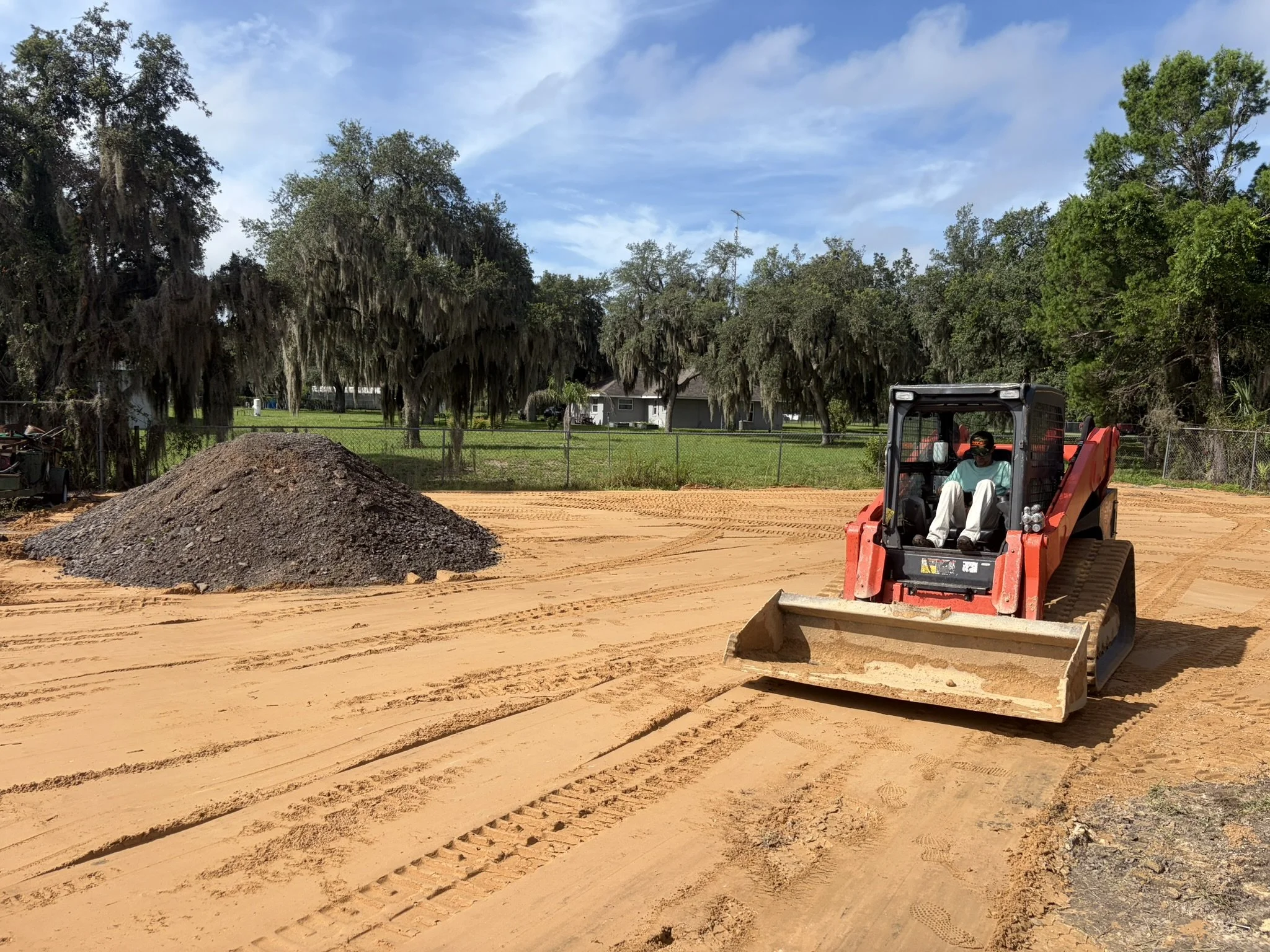 A man operating a small orange bulldozer on a construction site with orange dirt, surrounded by green trees, blue sky with some clouds, and a house in the background.