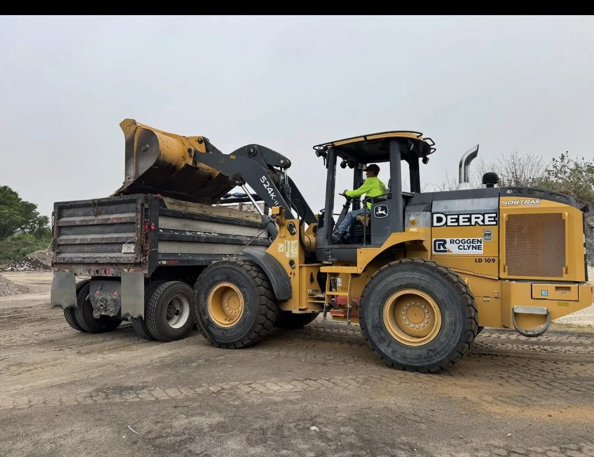 A yellow John Deere front loader with a bucket attachment, operated by a person in a neon green jacket, at a construction or dirt site.