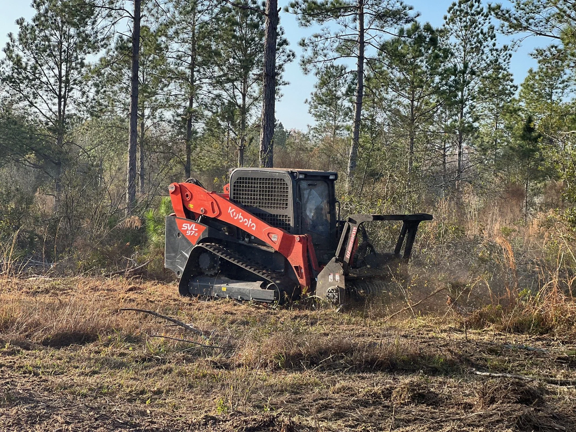 A small orange Kubota compact utility tractor with tracks instead of wheels, sitting on a cleared patch of land surrounded by trees.