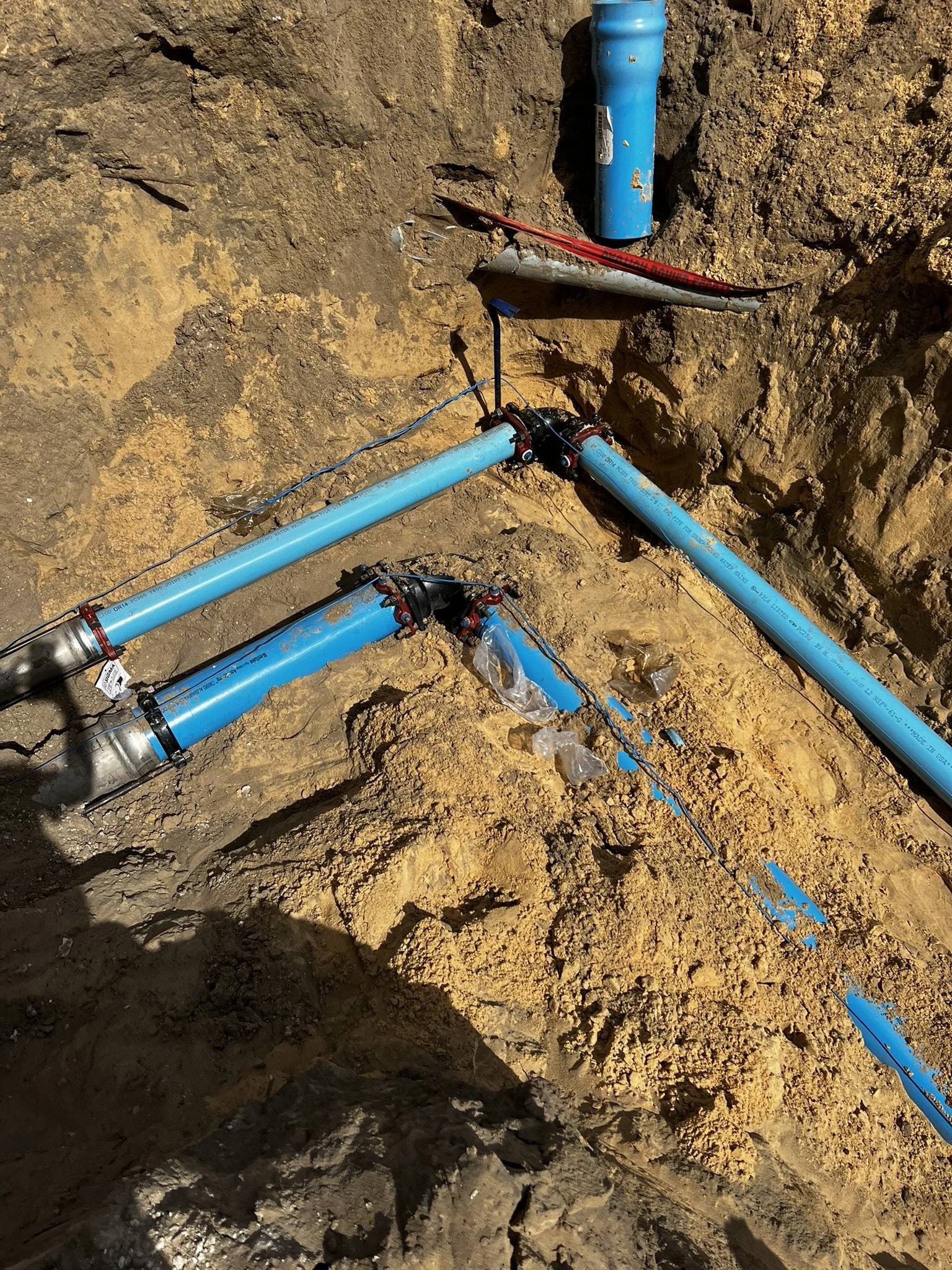 Blue underground water pipes installed in an excavated dirt trench, connected with fittings, with some wires attached, in a construction or repair site.