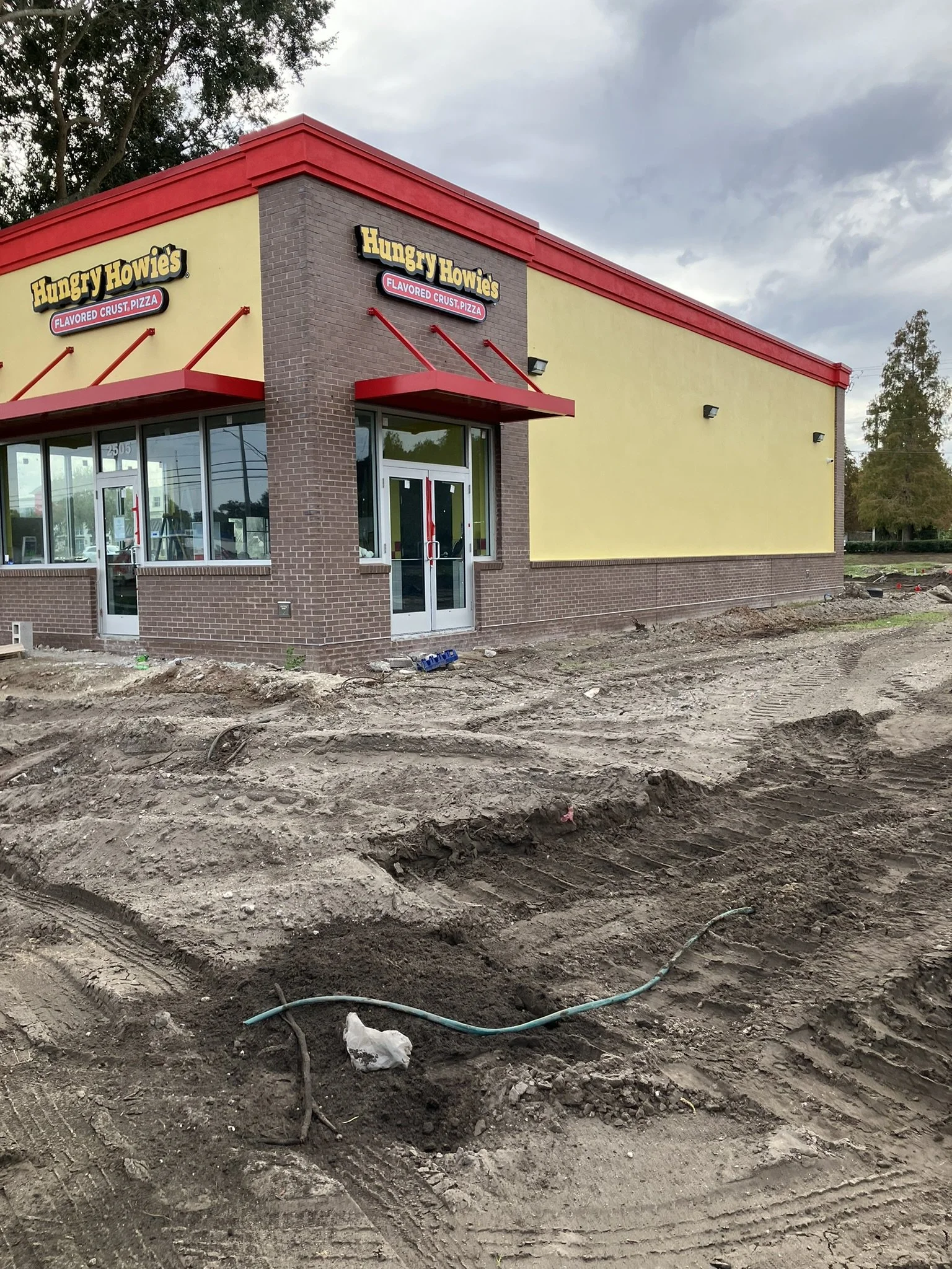 Construction site with dirt ground, tire tracks, and exposed pipes, in front of a fast food restaurant building with yellow walls, a red roof edge, and signage for Hungry Howie's pizza.