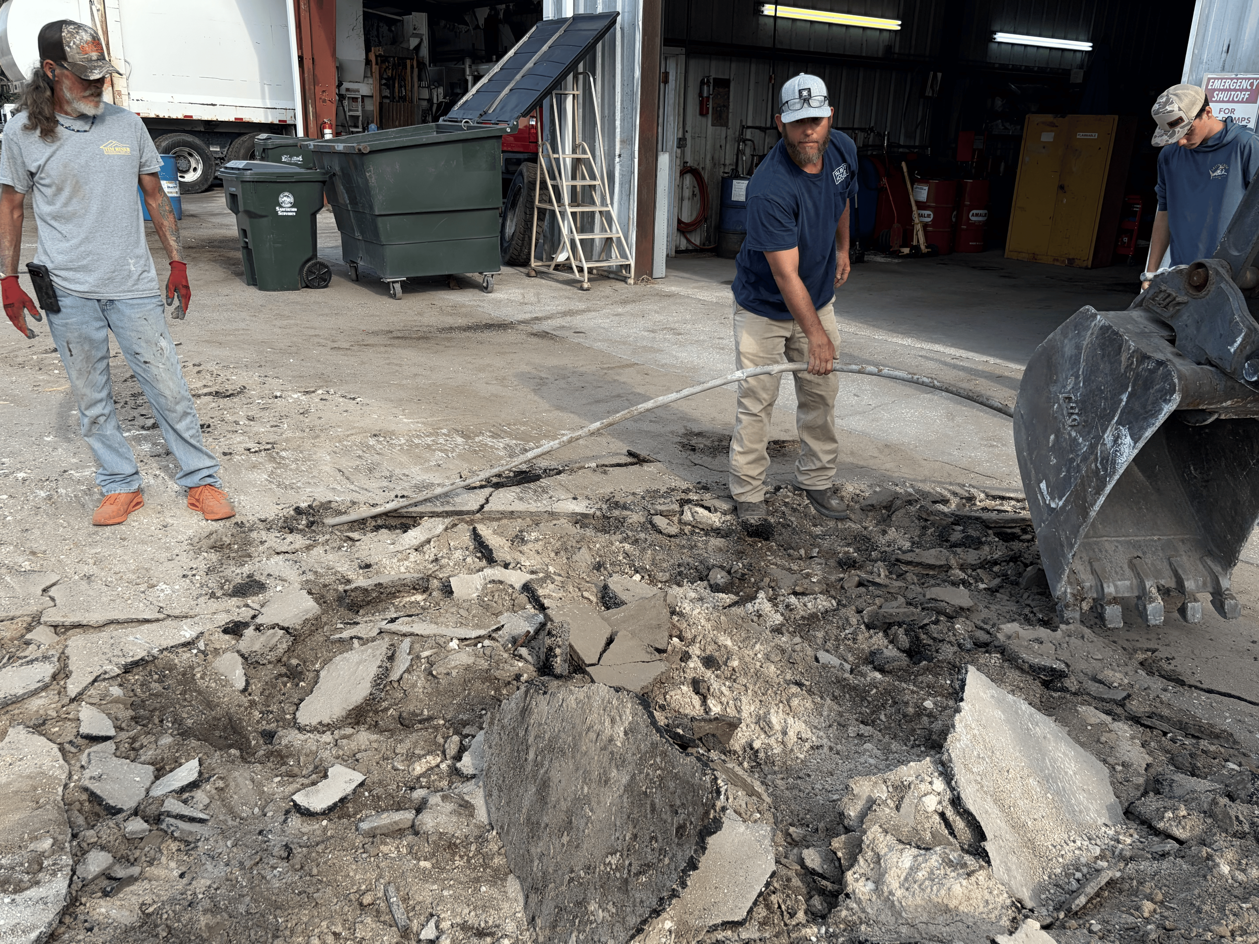 Workers repairing a collapsed section of asphalt on a driveway or parking lot, with one person holding a jackhammer and debris scattered around.