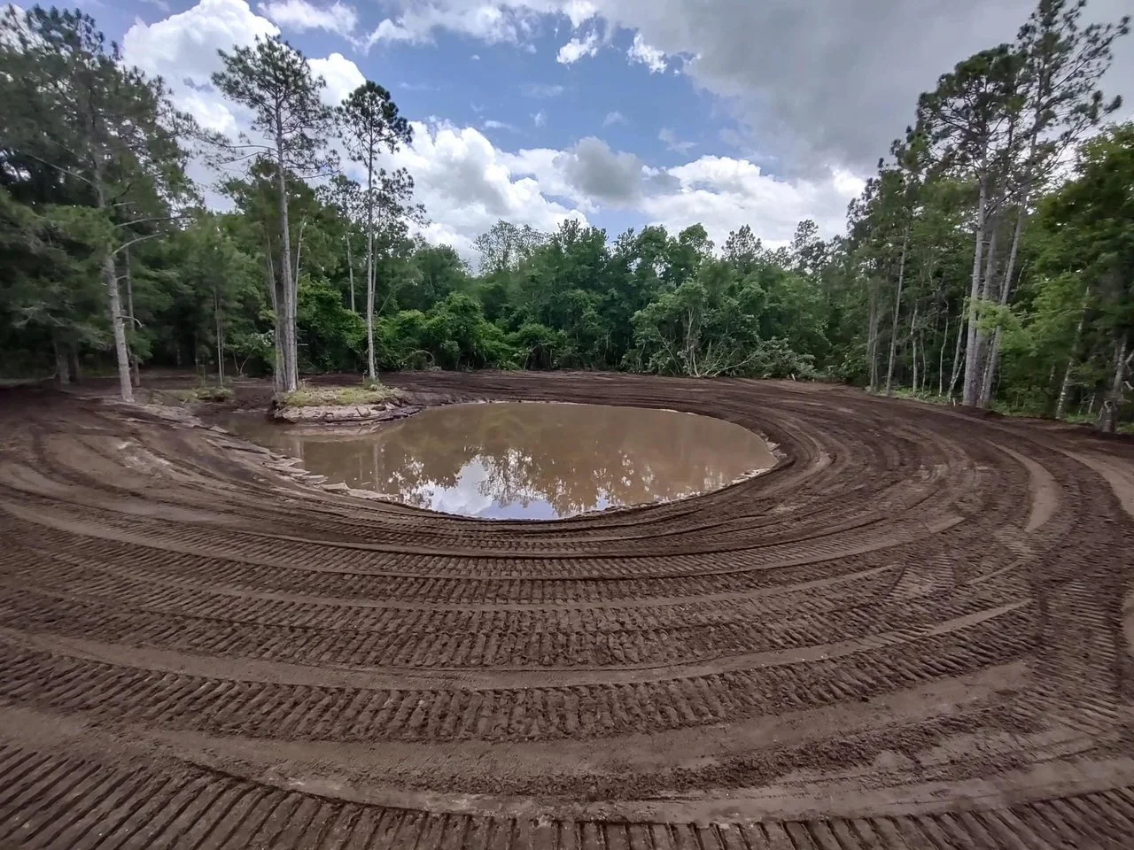 A small muddy pond surrounded by a dirt track with tire marks, situated in a forested area with green trees and partly cloudy sky.