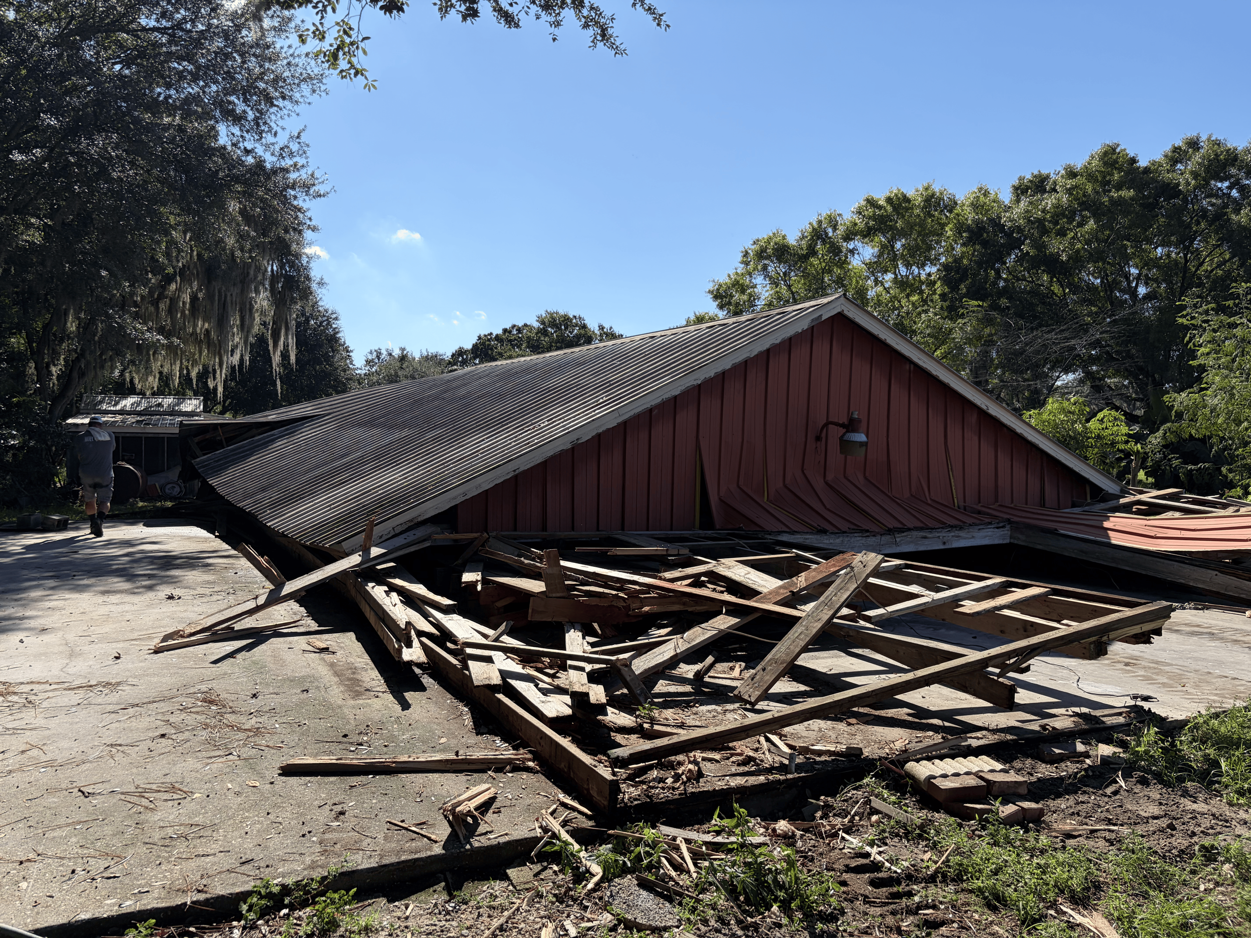 A red building with a metal roof that has collapsed, with debris and broken wood scattered on the ground. There are trees with green leaves around, and the sky is blue with a few clouds.
