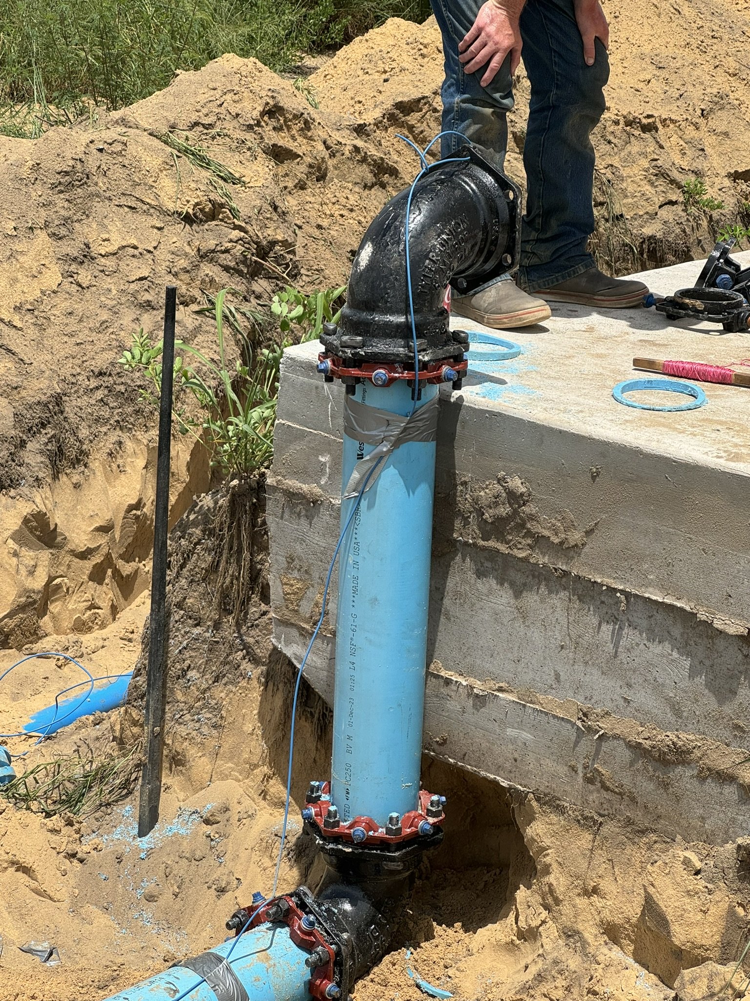 Plumbing installation with large black valve, blue pipe, and red fittings in a trench at a construction site.