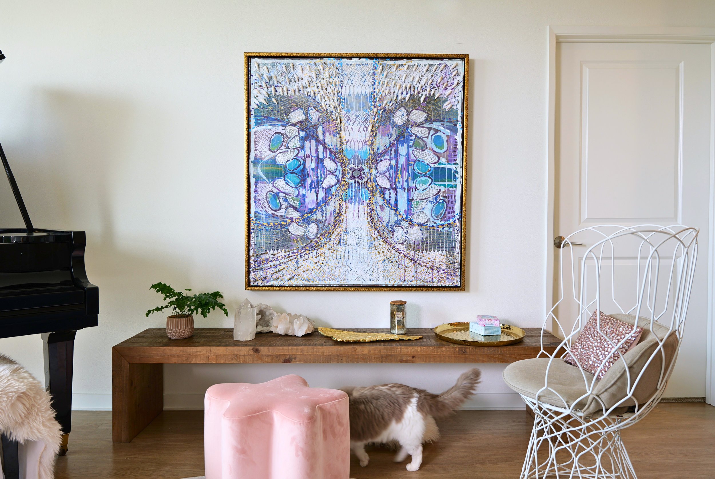 Interior of a living room with a large framed artwork of a butterfly on the wall, a wooden table with decorative objects, a small potted plant, a pink velvet stool, a white wire chair, and a fluffy cat walking underneath it.