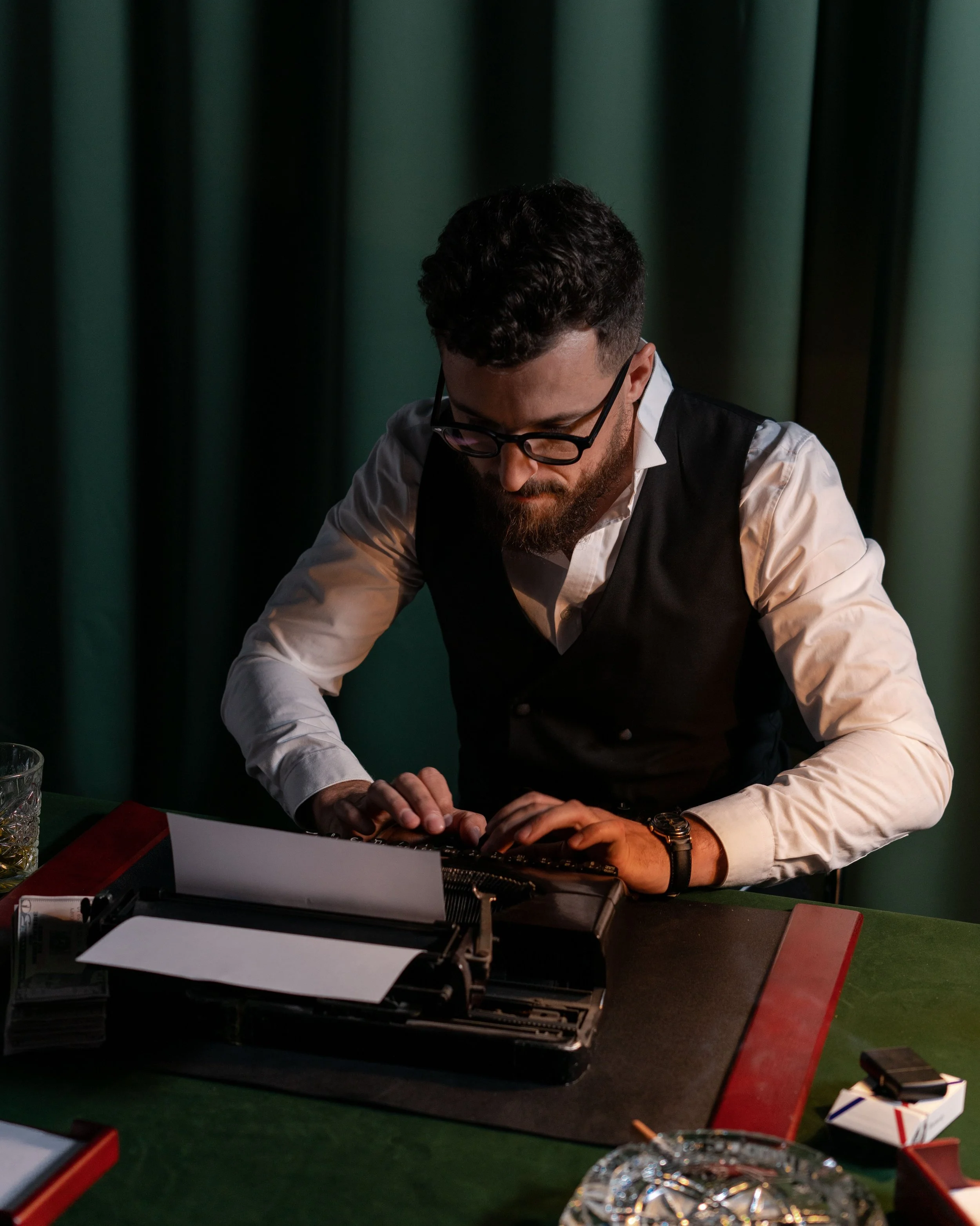 Man with dark curly hair and beard, wearing glasses, white shirt, and black vest, sitting at a table with a typewriter, in front of green curtain.