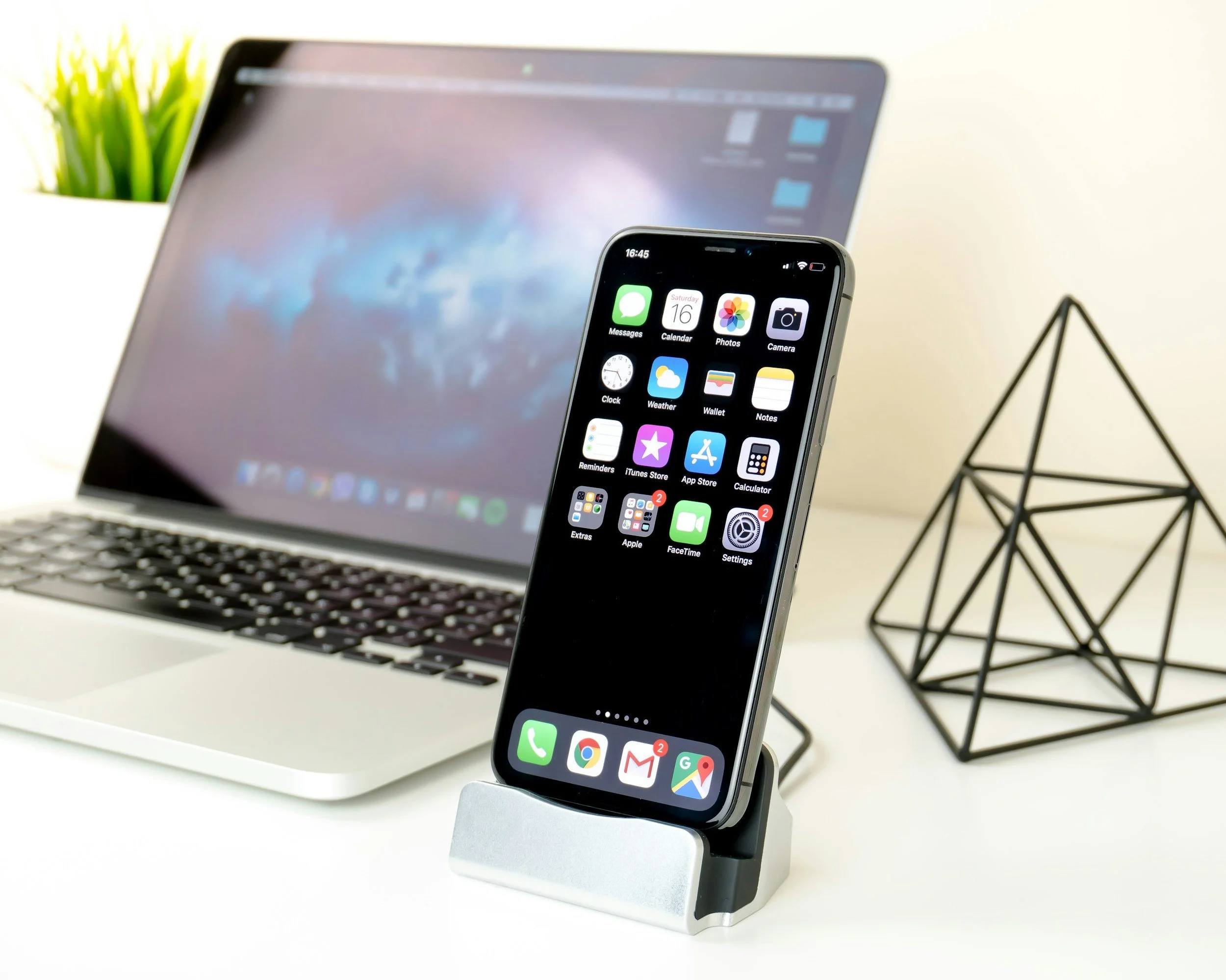 A smartphone displaying various app icons is placed on a silver stand on a white desk. Behind the phone, there is a laptop with a blurred screen and a potted plant with green leaves. A black wireframe decorative object is also visible on the right side of the desk.