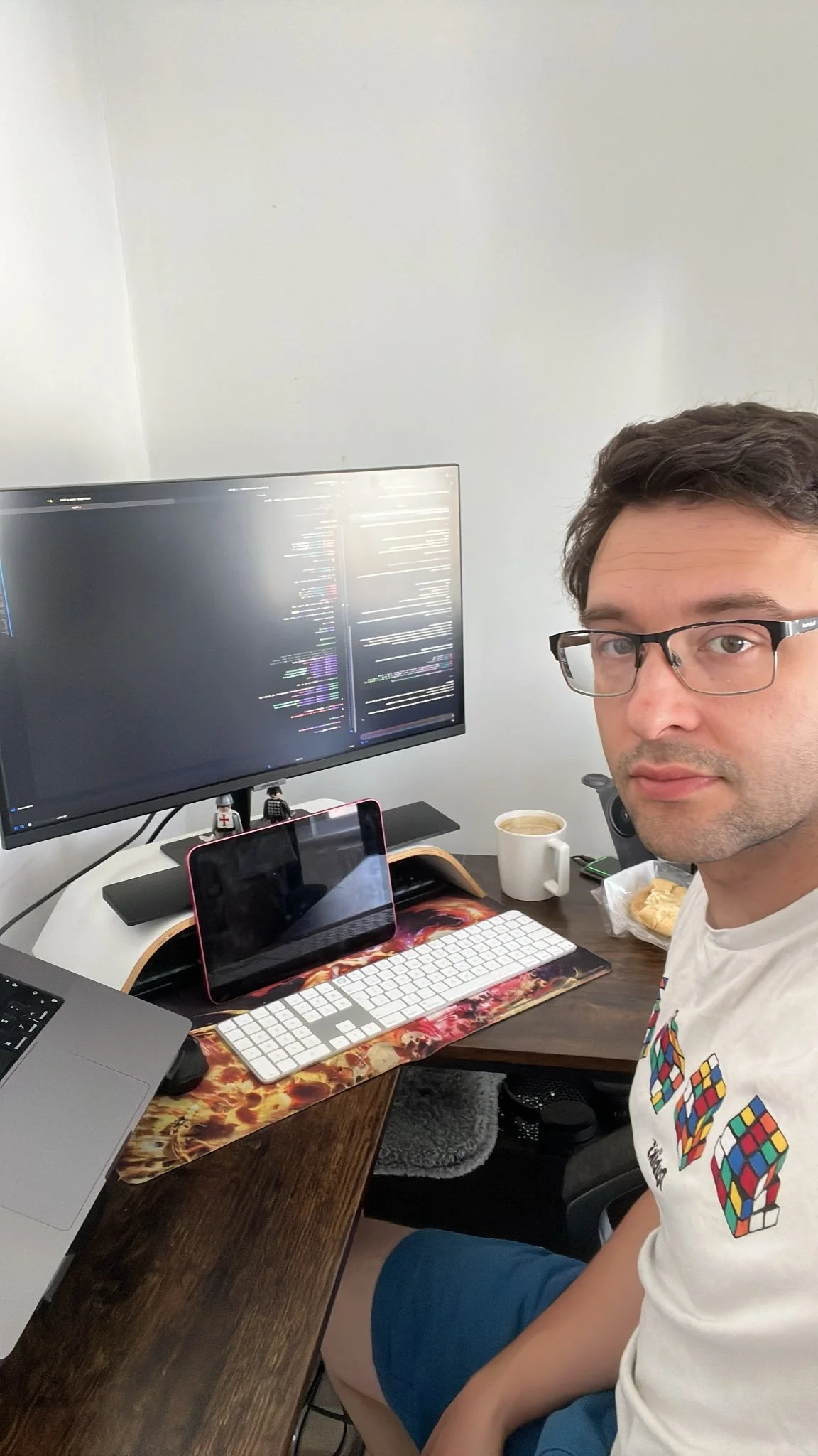 A person with glasses taking a selfie at a work desk with dual monitors displaying codes, a coffee mug, a keyboard, a mouse, an iPad, and some snacks.