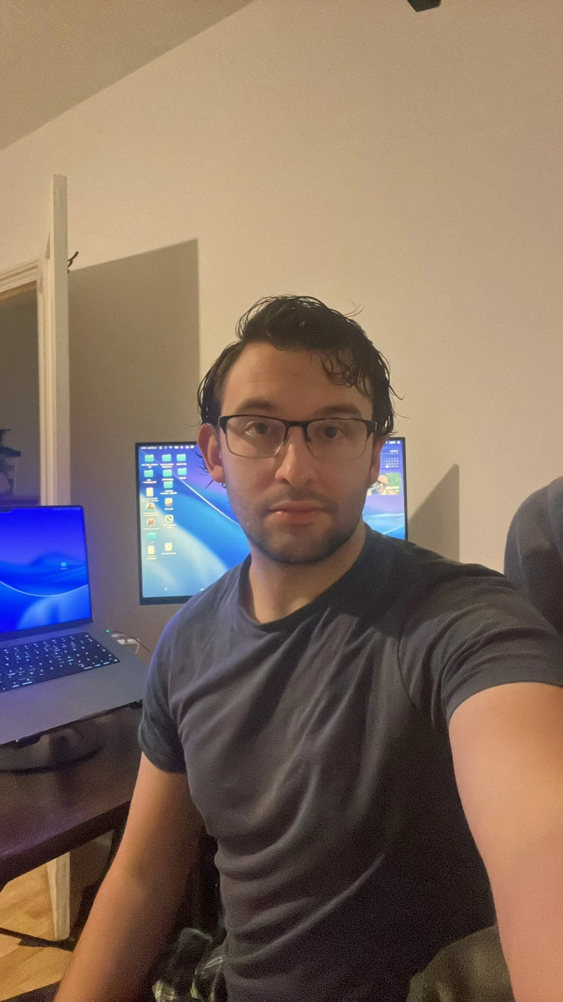 A young man with dark hair and glasses taking a selfie in a room with two computer monitors on a desk behind him.