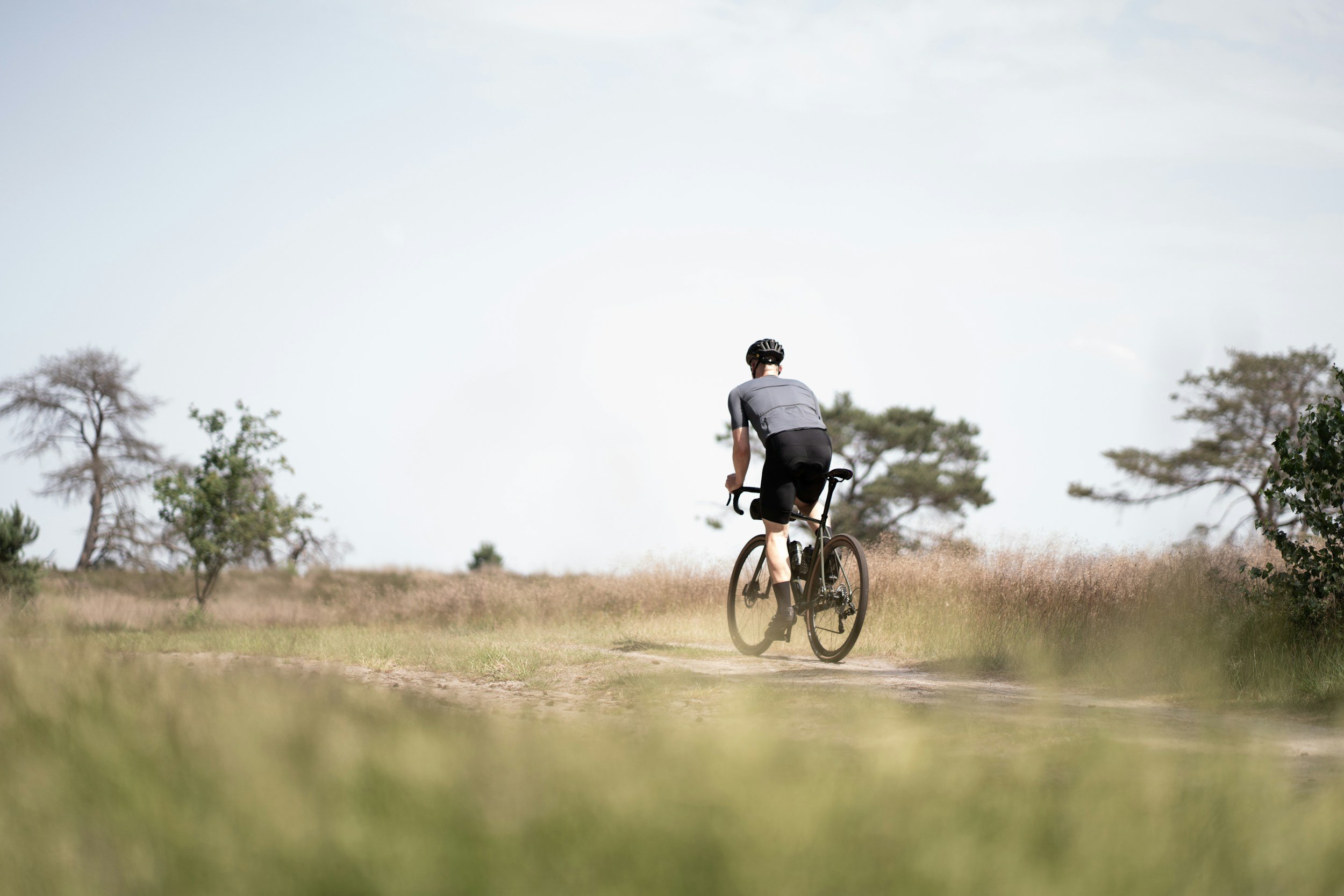 A person riding a bicycle on a dirt path in a park with trees and grass, under a partly cloudy sky.