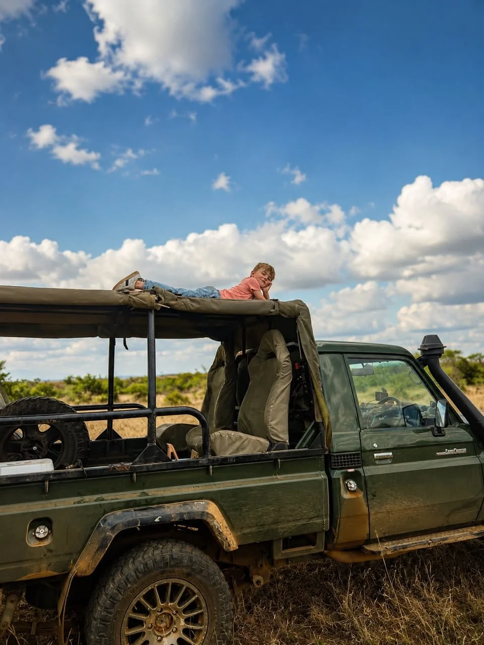 A young girl is lying on top of a green off-road vehicle with a lifted roof, in a grassy field under a partly cloudy sky.