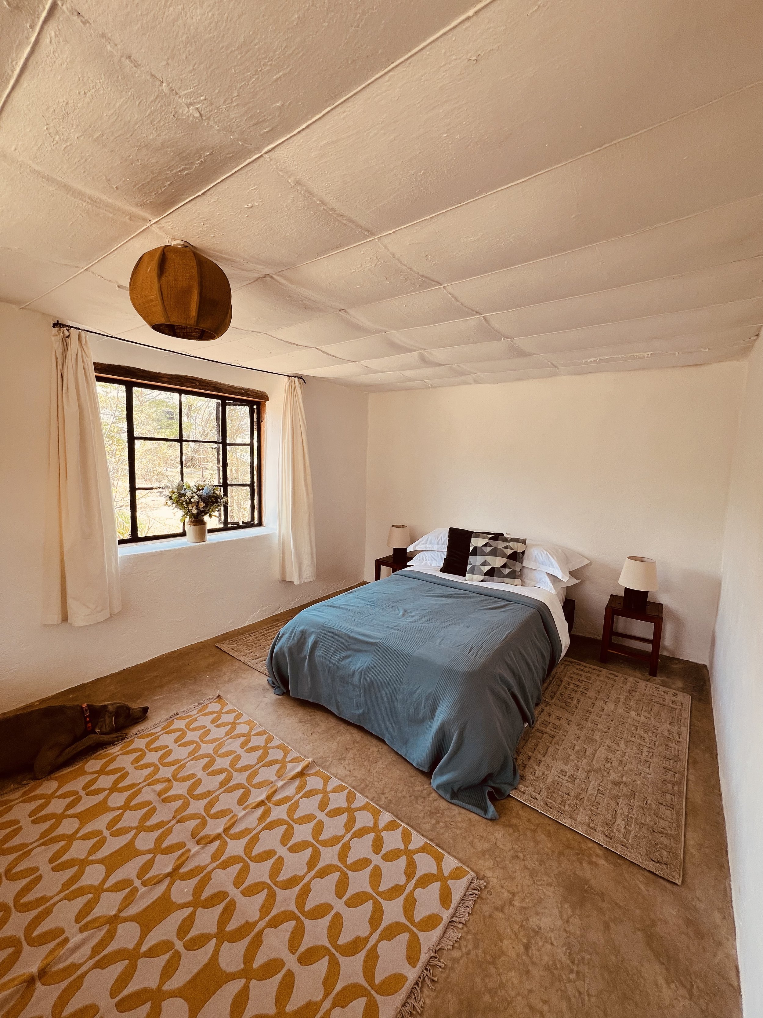 A bedroom with a bed, window with curtains, and two nightstands with lamps. The ceiling appears textured and has a hanging light fixture. A dog is lying on a colorful patterned rug in the corner of the room.
