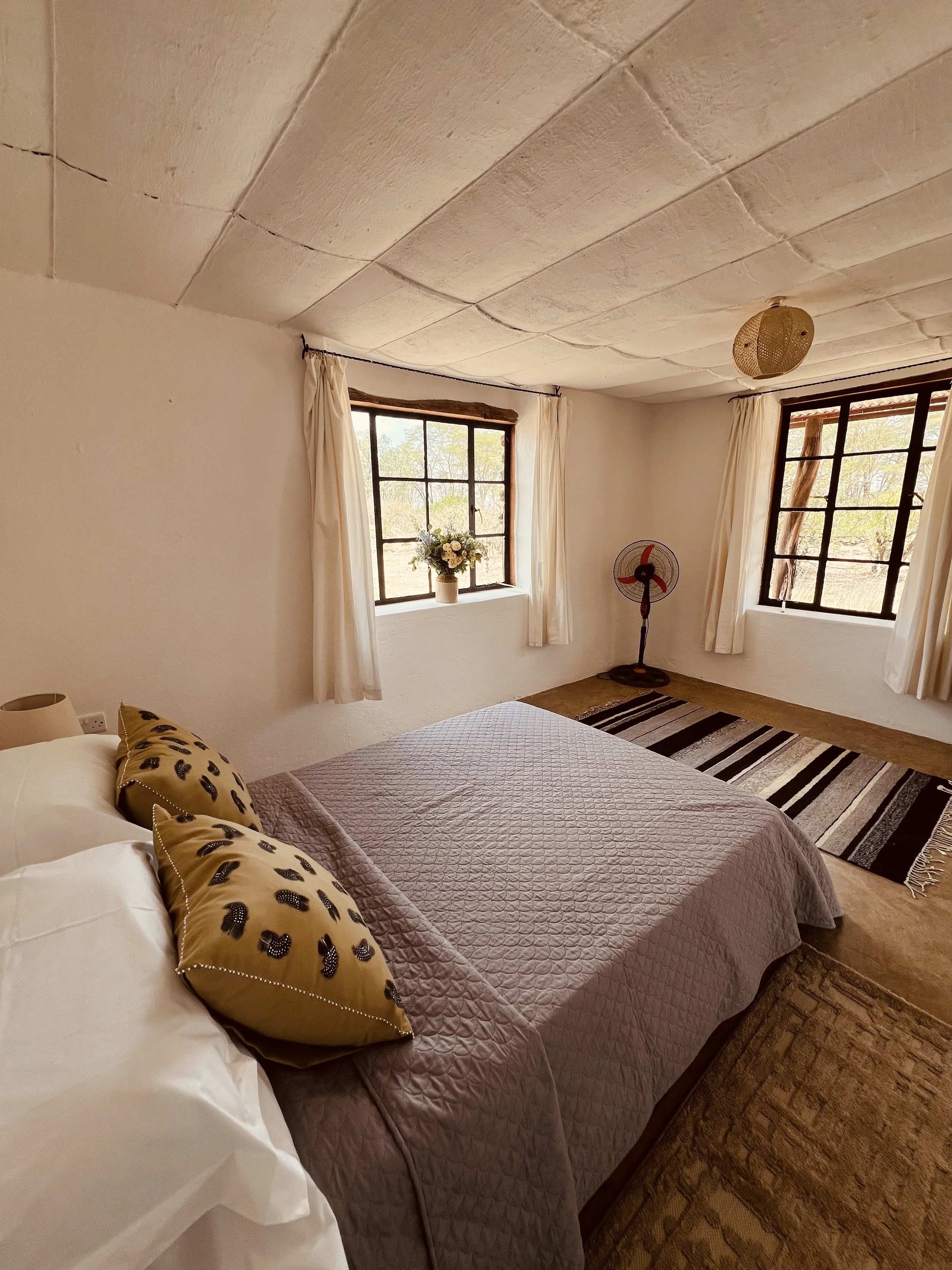 A cozy bedroom with a large bed, tan and black decorative pillows, and a pink quilted bedspread. There are two windows with cream curtains, a black and white striped rug, a standing fan, and a vase with flowers on the windowsill. The ceiling appears to be made of panels or tiles, and there is a woven light fixture hanging from it.