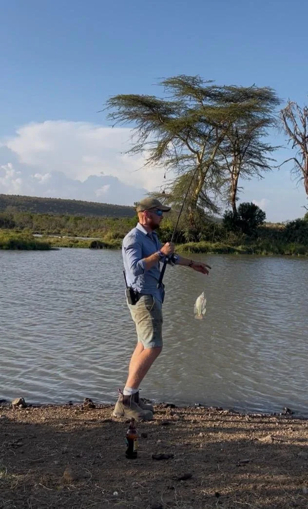 Man standing by a lake fishing with a rod, holding a fish he caught, with trees and a blue sky in the background.