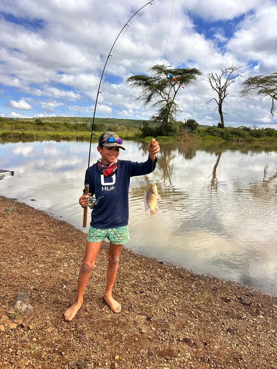 A young boy wearing a hat, sunglasses, a navy hoodie, and green shorts with a fish caught on his fishing line, standing barefoot near a lake with trees and cloudy sky in the background.