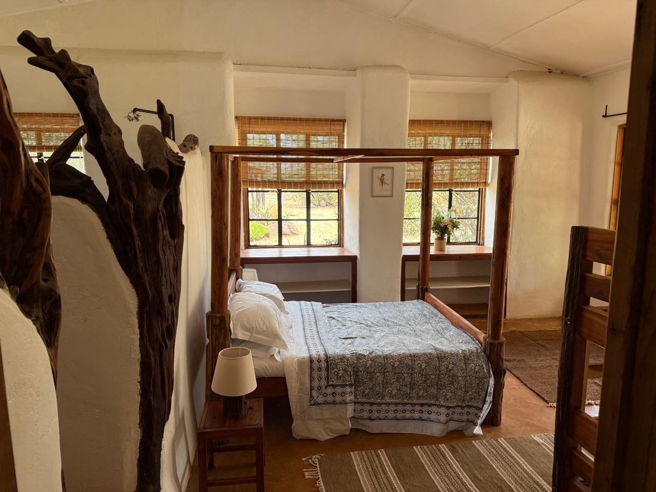 Cozy bedroom with a wooden four-poster bed, a nightstand with a lamp, and three large windows with bamboo shades, decorated with a framed artwork and potted plants.