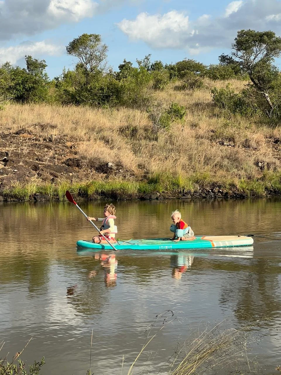 Two children on a paddleboard in a calm river, with one child paddling and the other sitting, surrounded by grassy hills and trees under a partly cloudy sky.