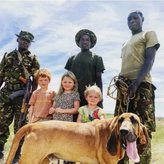 Three soldiers in camouflage and green shirts stand with three young girls and a large dog outdoors under a blue sky.