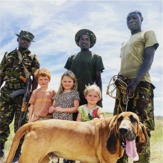 Three children and three soldiers in a field, with a large dog in the foreground, under a cloudy sky.
