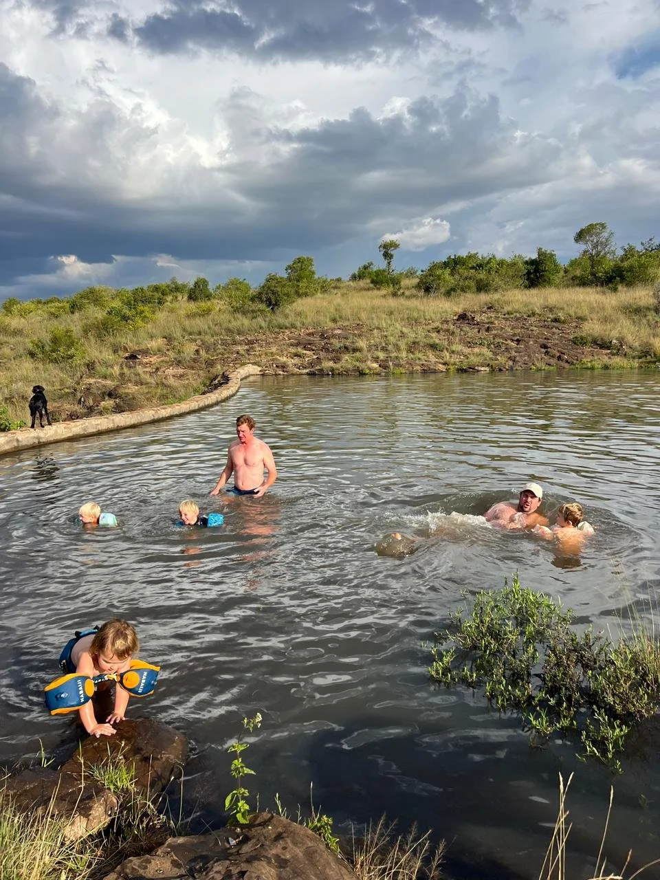 A group of people swimming and playing in a natural body of water outdoors, with a grassy hill and cloudy sky in the background.