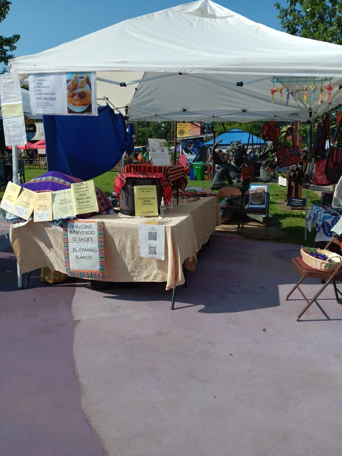 A market stall at an outdoor fair with a white canopy tent selling handmade crafts and textiles. There are signs with prices and descriptions, colorful bags, and a statue displayed behind the table. The sky is clear and trees are visible in the background.