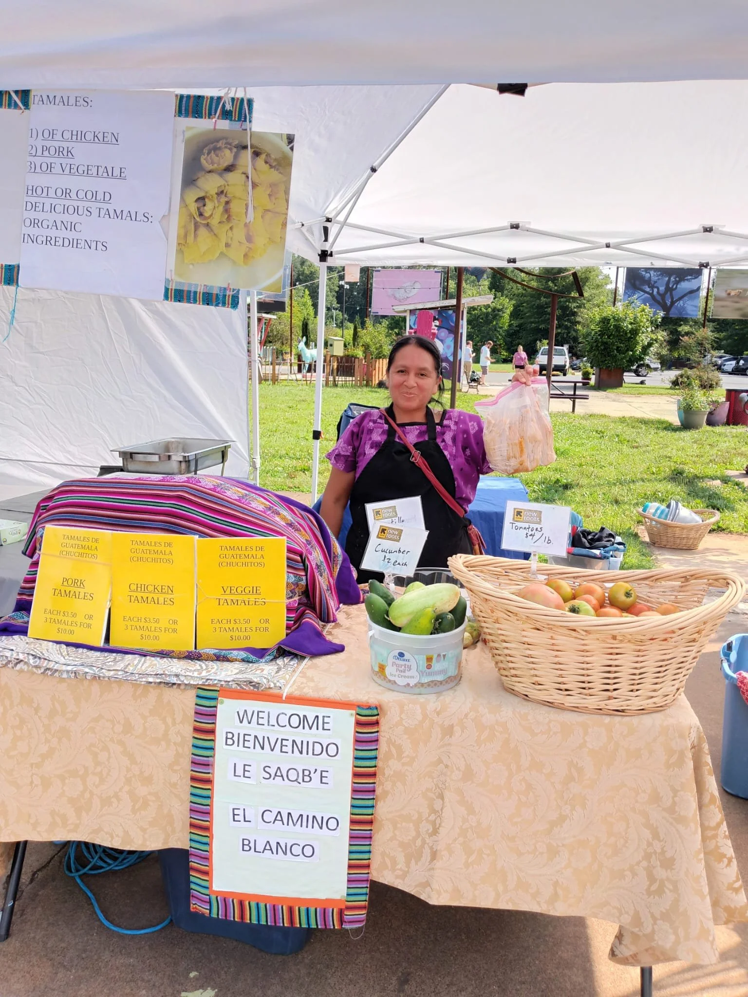 A woman standing behind a table at a market stall with various signboards and baskets of fresh produce. The table is covered with a floral tablecloth and a welcome sign with bilingual text. The background shows trees, a park, and a few people walking.