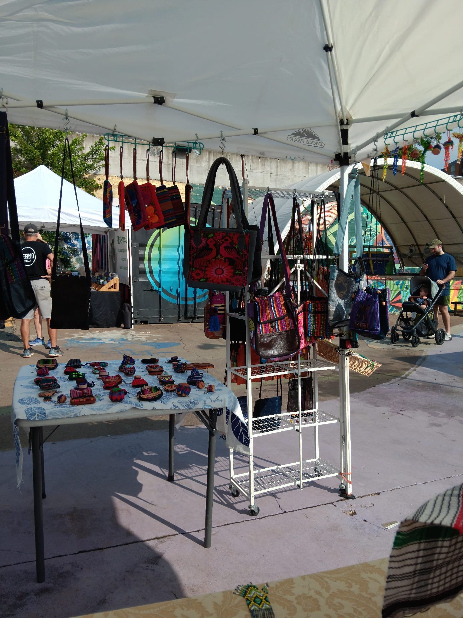 A market stall displayed under a white canopy, featuring colorful woven bags and accessories, with people browsing in the background at an outdoor event.