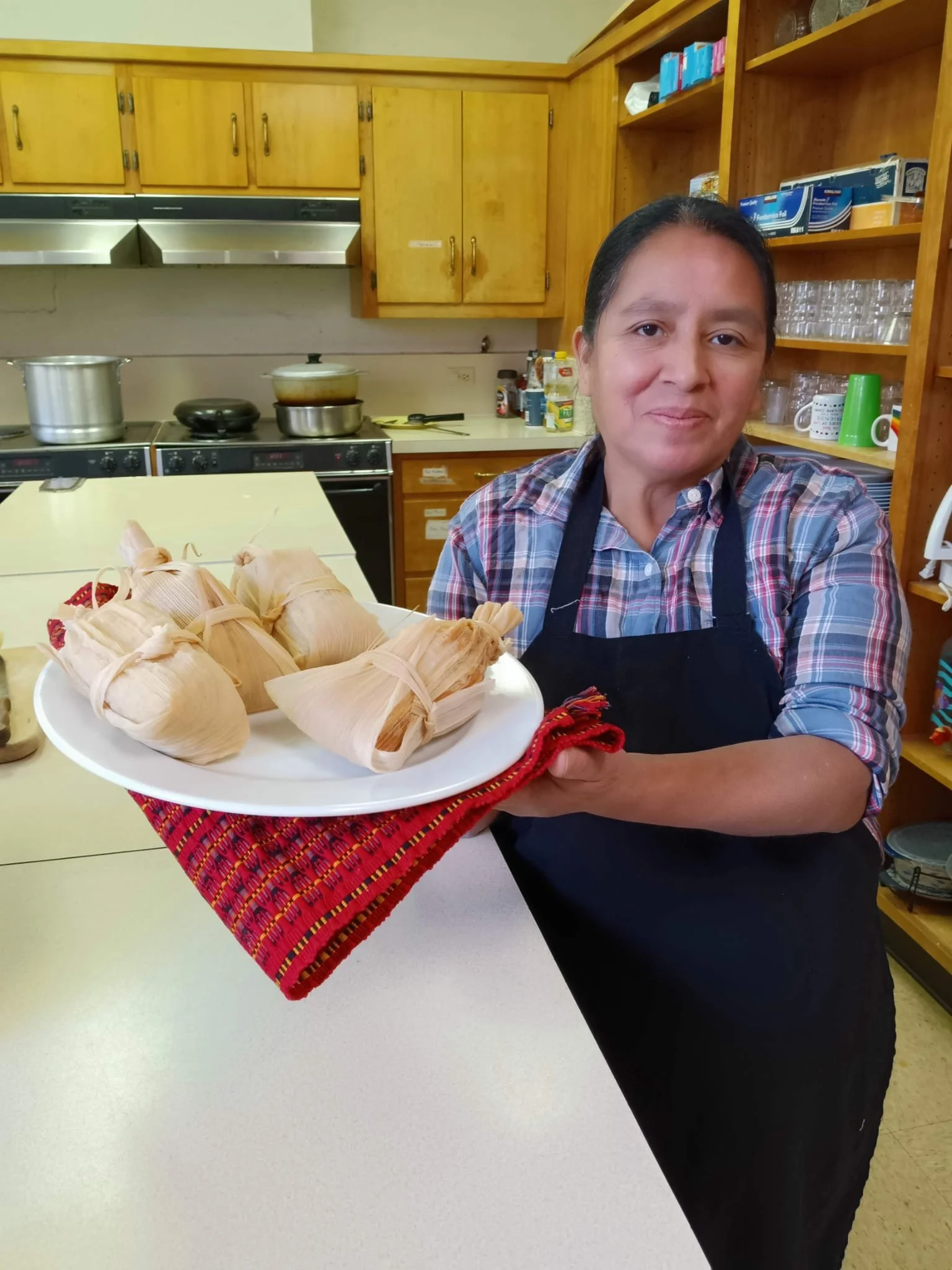A woman in a plaid shirt and apron holding a plate with several tamales wrapped in corn husks, standing in a kitchen.