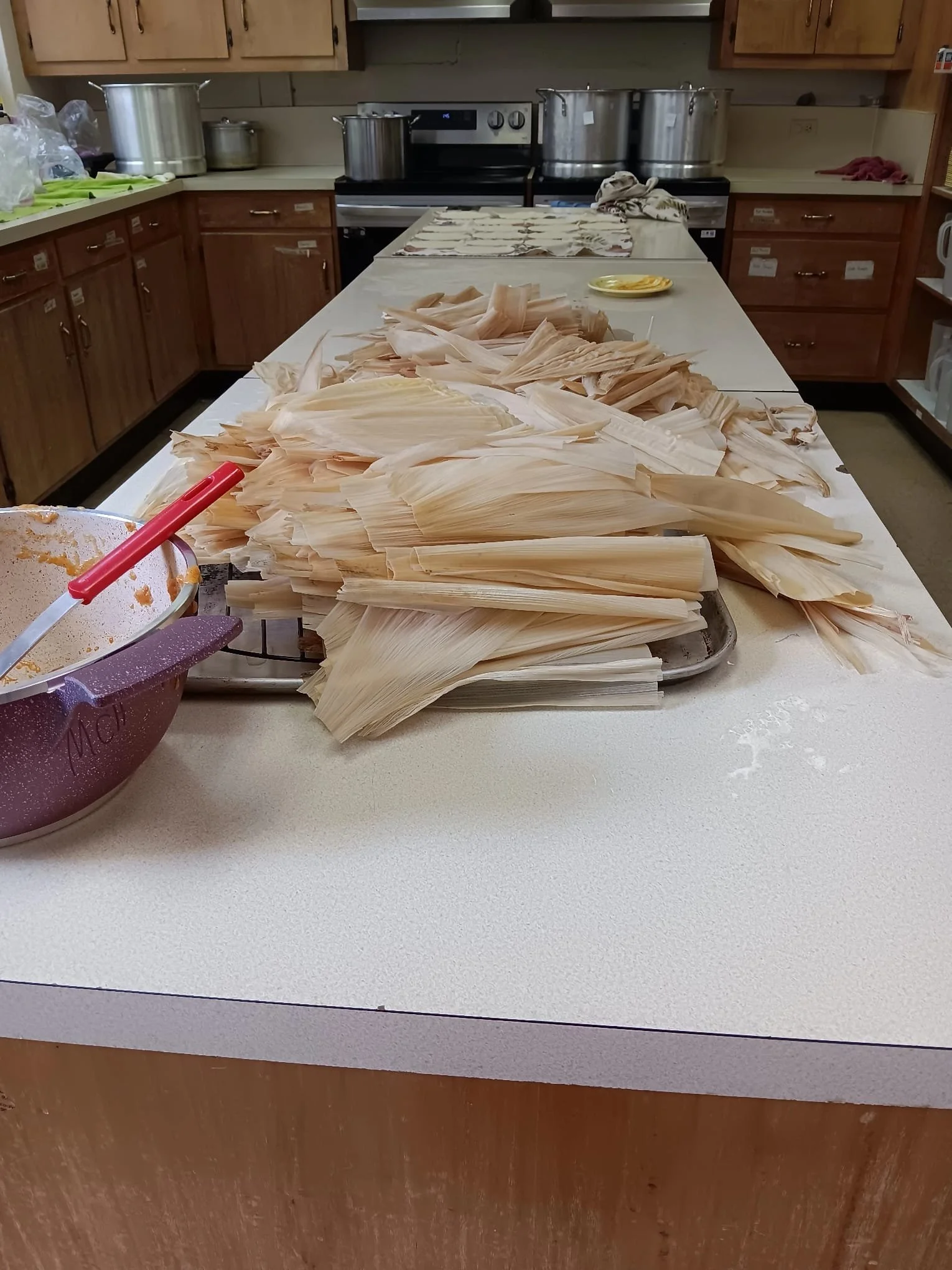 A kitchen with a white countertop, wooden cabinets, and several large stainless steel pots on the stove. On the countertop, there are piles of dried corn husks, a purple bowl with a red spoon, and a yellow plate.
