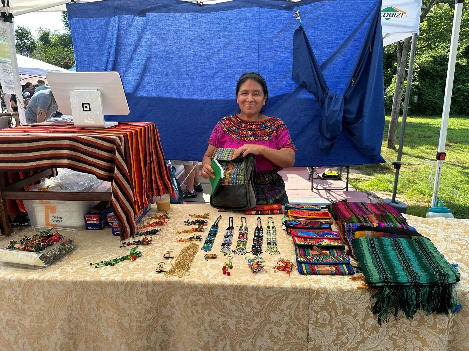 Woman standing behind a table at an outdoor market, displaying colorful handmade jewelry, textiles, and accessories.