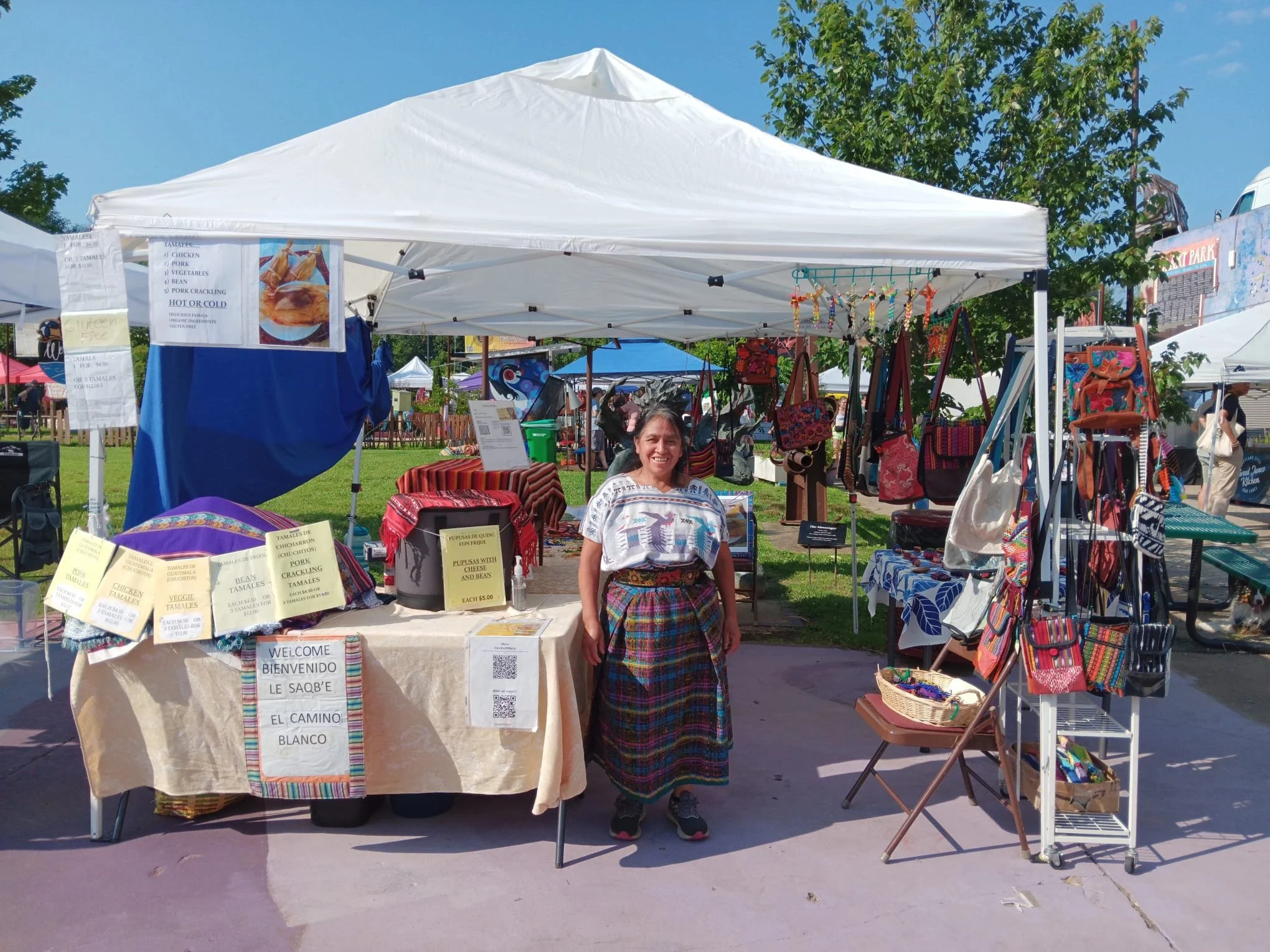 A woman sitting in front of a booth at an outdoor market, selling colorful handwoven textiles and crafts, with signs displaying prices and items in Spanish, under a white canopy on a sunny day.