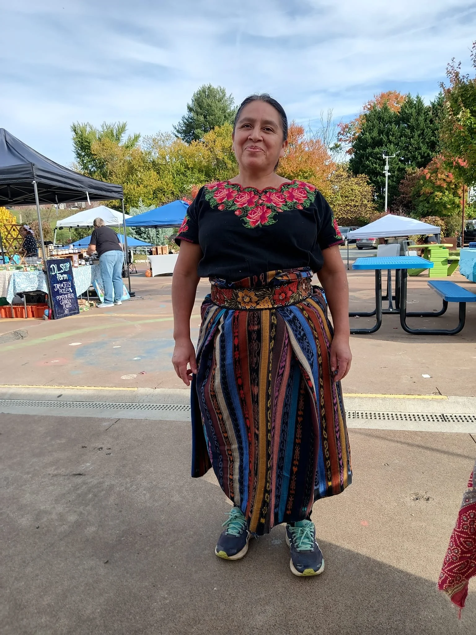 Woman standing outdoors at farmer's market. She is wearing a black embroidered blouse and a colorful striped skirt, with teal sneakers. There are tents and other people in the background, trees with fall foliage, and a blue sky.