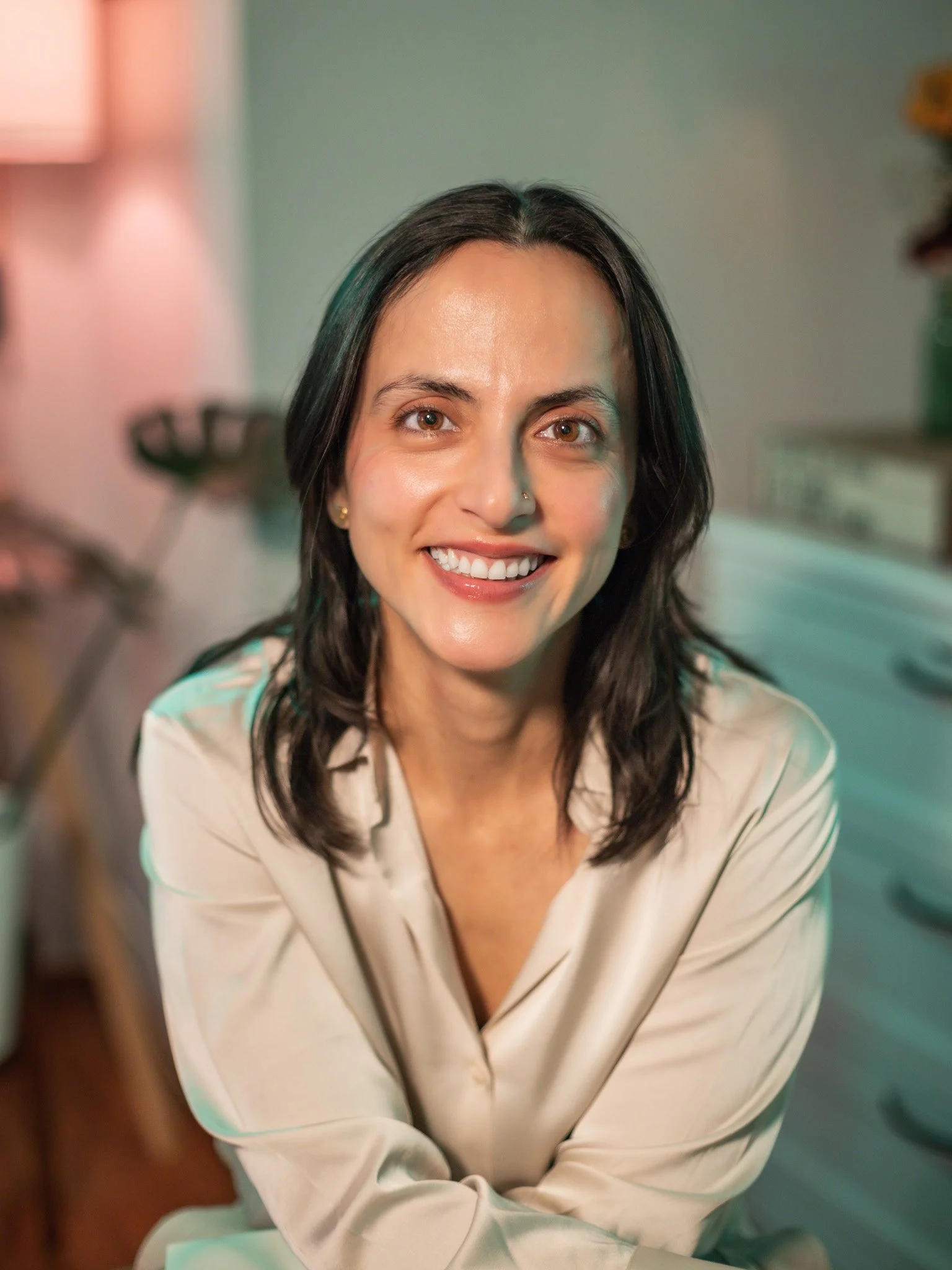 Close-up of a woman with shoulder-length dark hair smiling and looking at the camera, wearing a light-colored striped blazer, in a room with soft lighting.