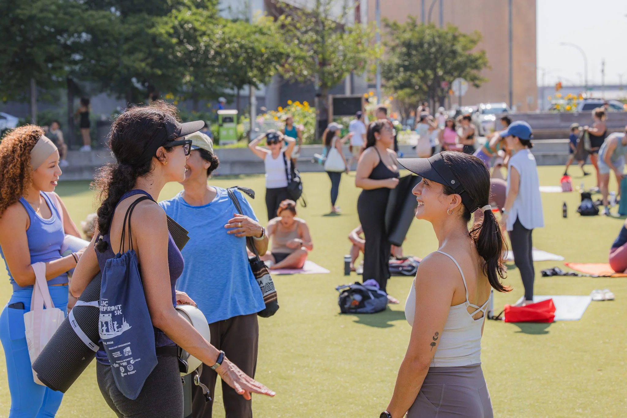 Two women in athletic clothing and visors are smiling and talking to each other in the foreground of a park. In the background, multiple people are engaging in fitness activities such as stretching and jogging on the grass. Trees and buildings are visible in the distance on a sunny day.