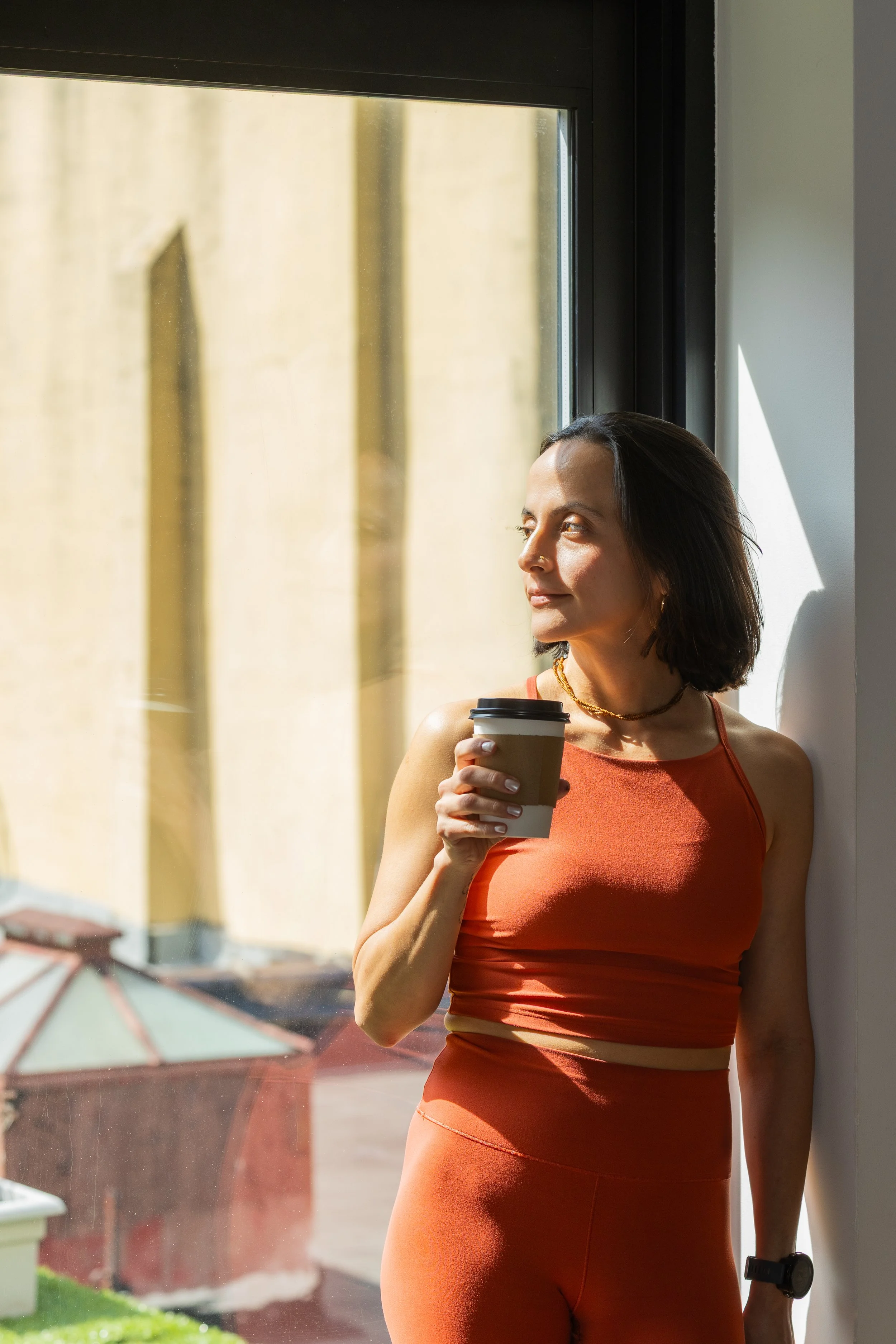 Woman in orange workout clothing holding a coffee cup, standing by a window, looking outside on a sunny day.