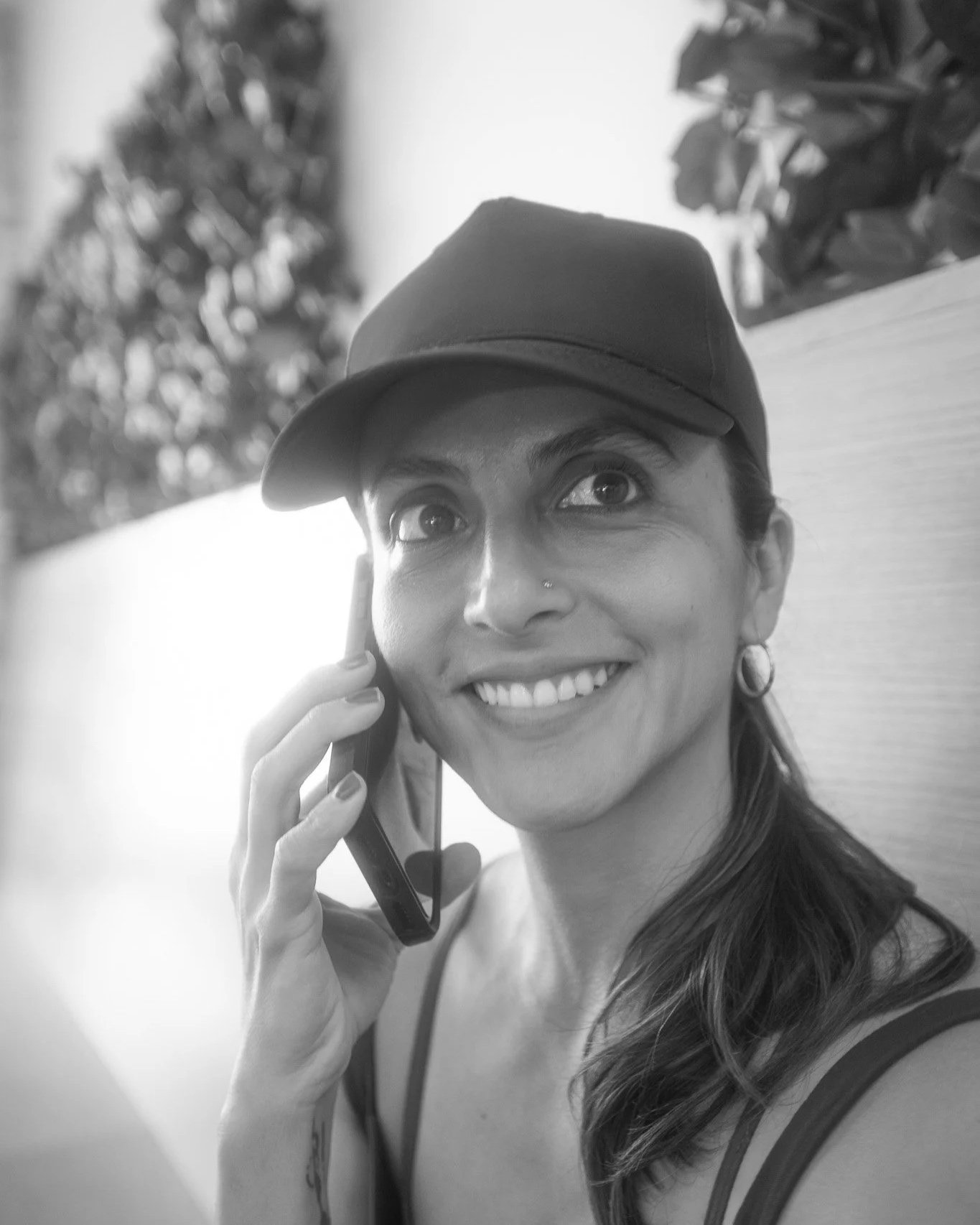 Black and white photo of a woman smiling and talking on a mobile phone, wearing a cap and earrings, with a blurred background of trees or bushes.