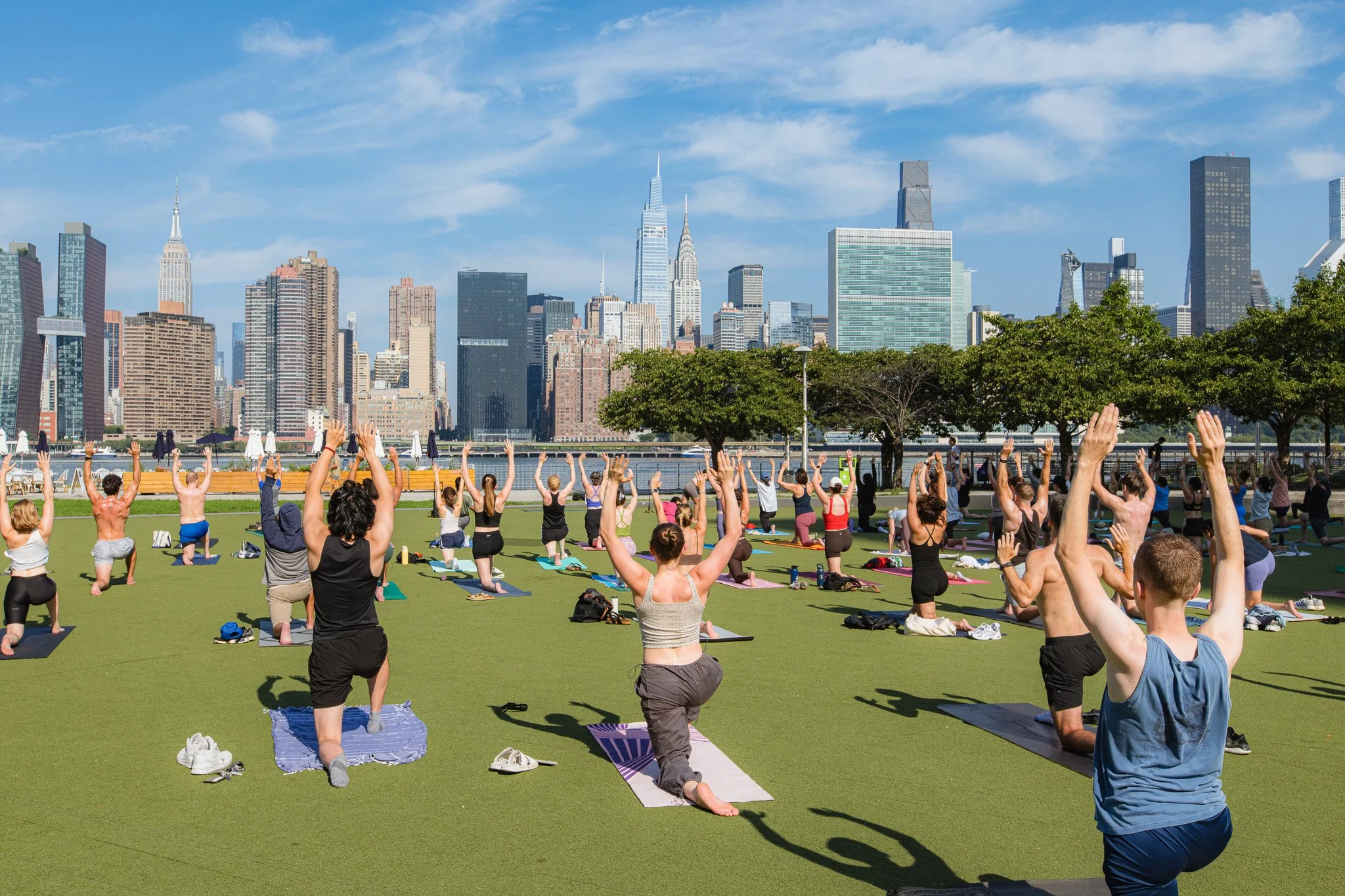 Group of people practicing yoga outdoors on a green field in front of a city skyline on a sunny day.